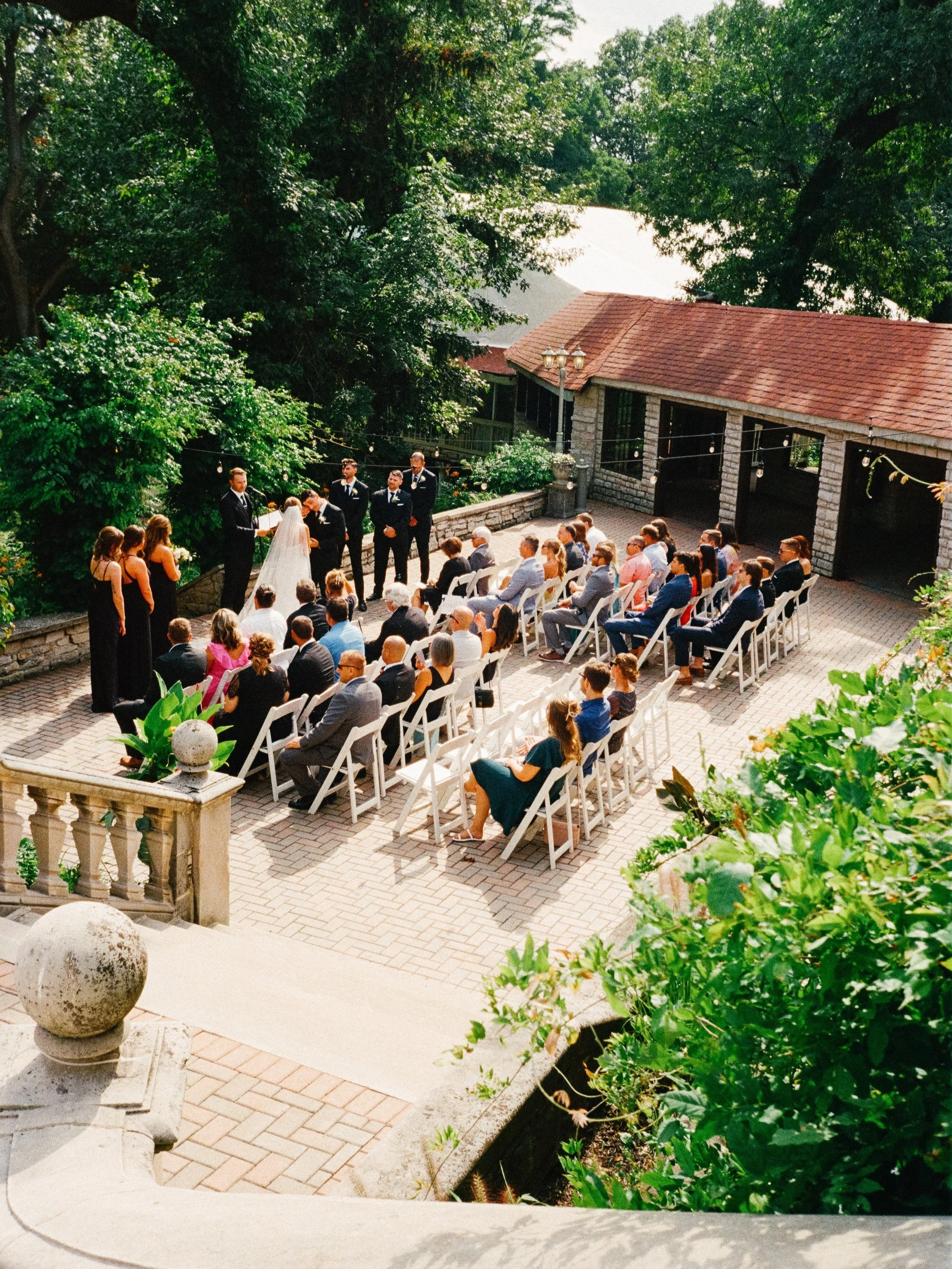 Outdoor wedding ceremony with guests seated in white chairs, officiant and bride and groom under trees, sunny day with string lights.