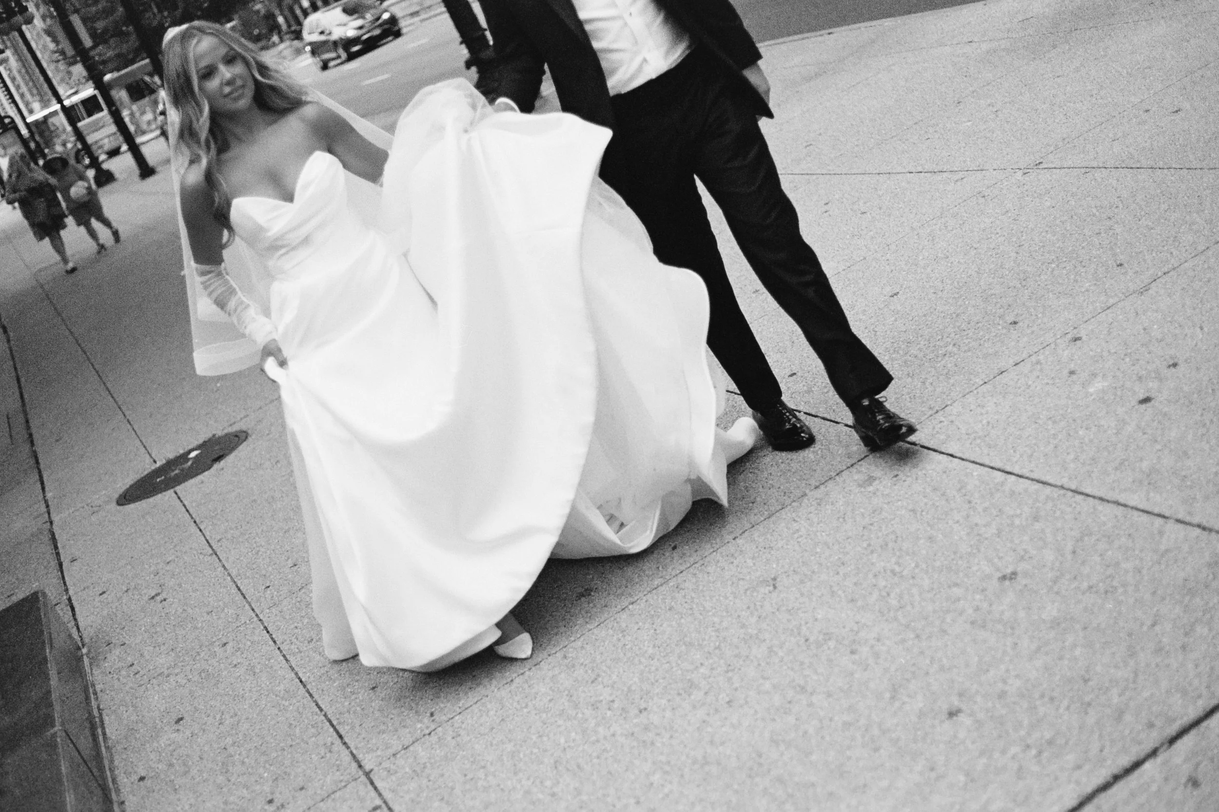 A woman in a wedding dress is being assisted by a man on a city sidewalk.
