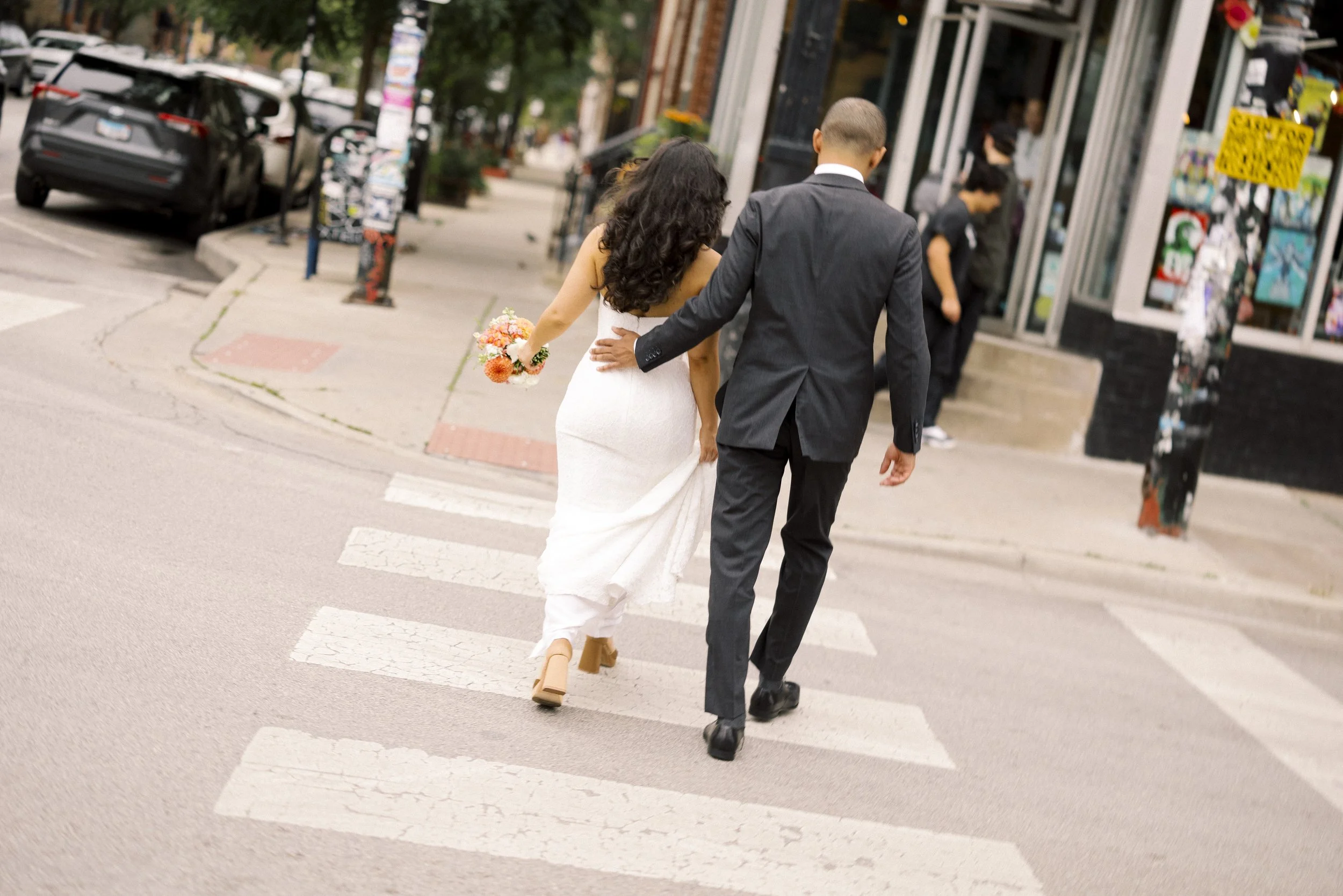 A newlywed couple crossing the street in an urban area, with the bride holding a bouquet and wearing a white wedding dress and the groom in a dark suit, walking hand in hand.