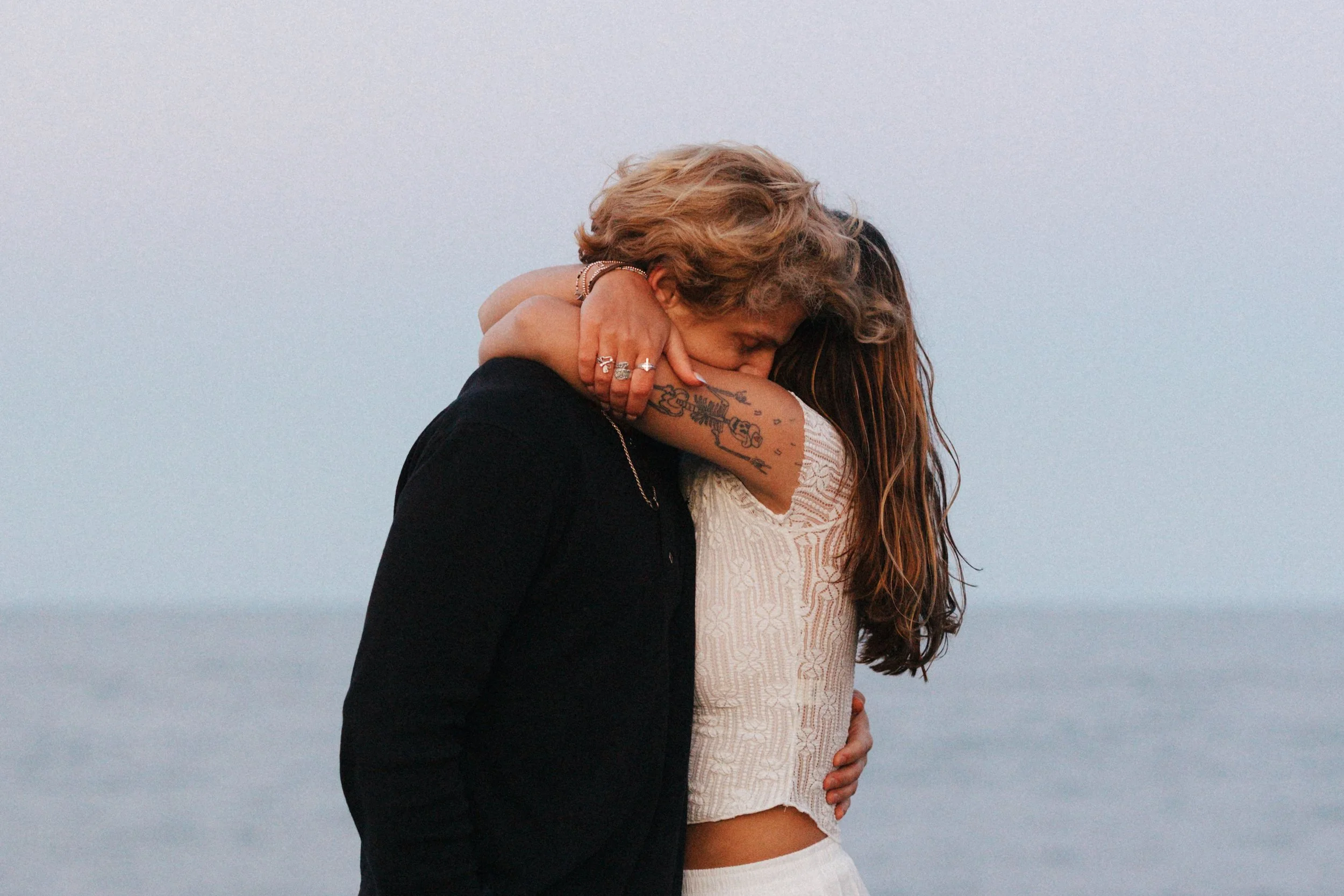 Two women embracing each other closely, with a body of water and a clear sky in the background.