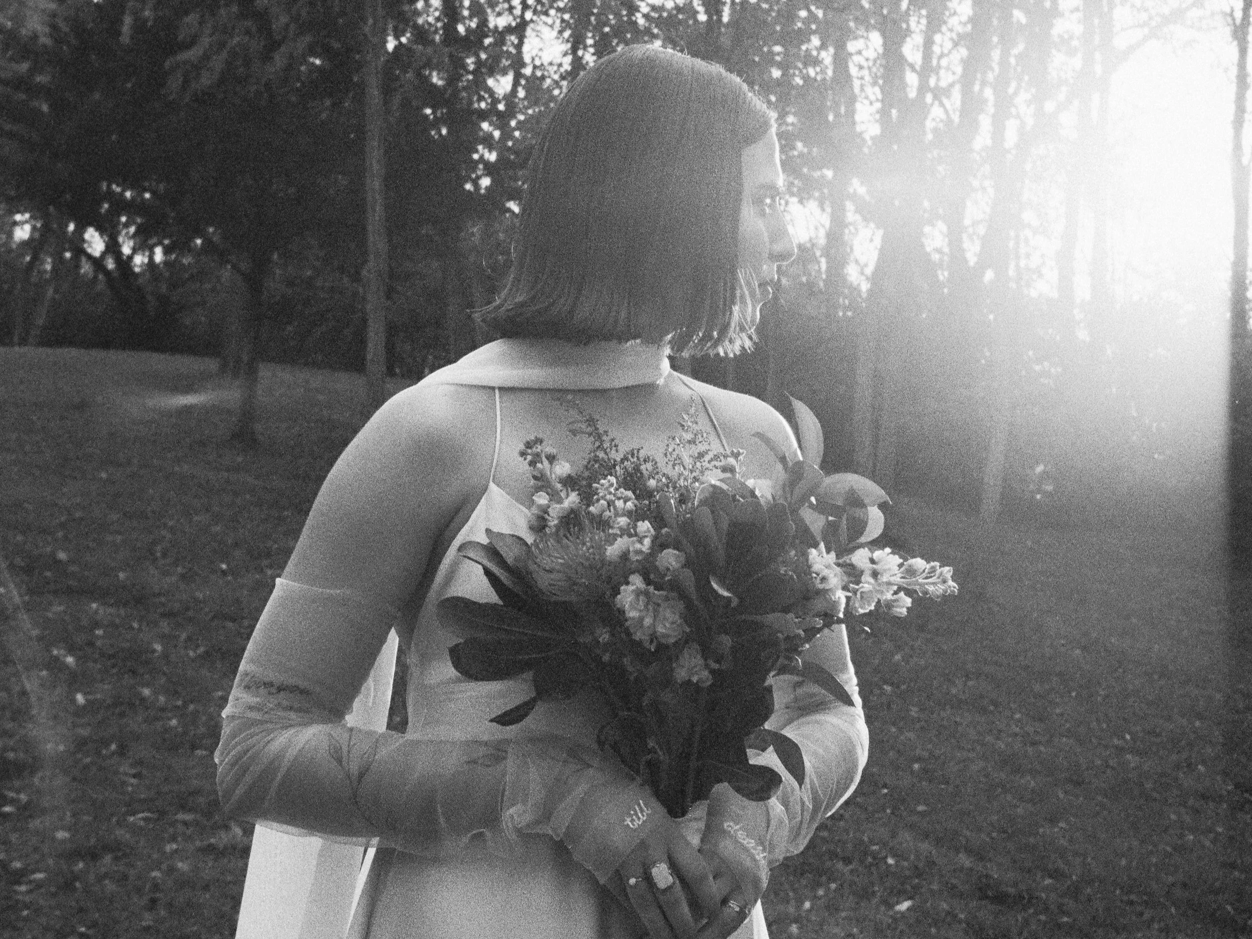 A woman in bridal attire holding a bouquet of flowers, standing outdoors with trees in the background and sunlight backlighting the scene.