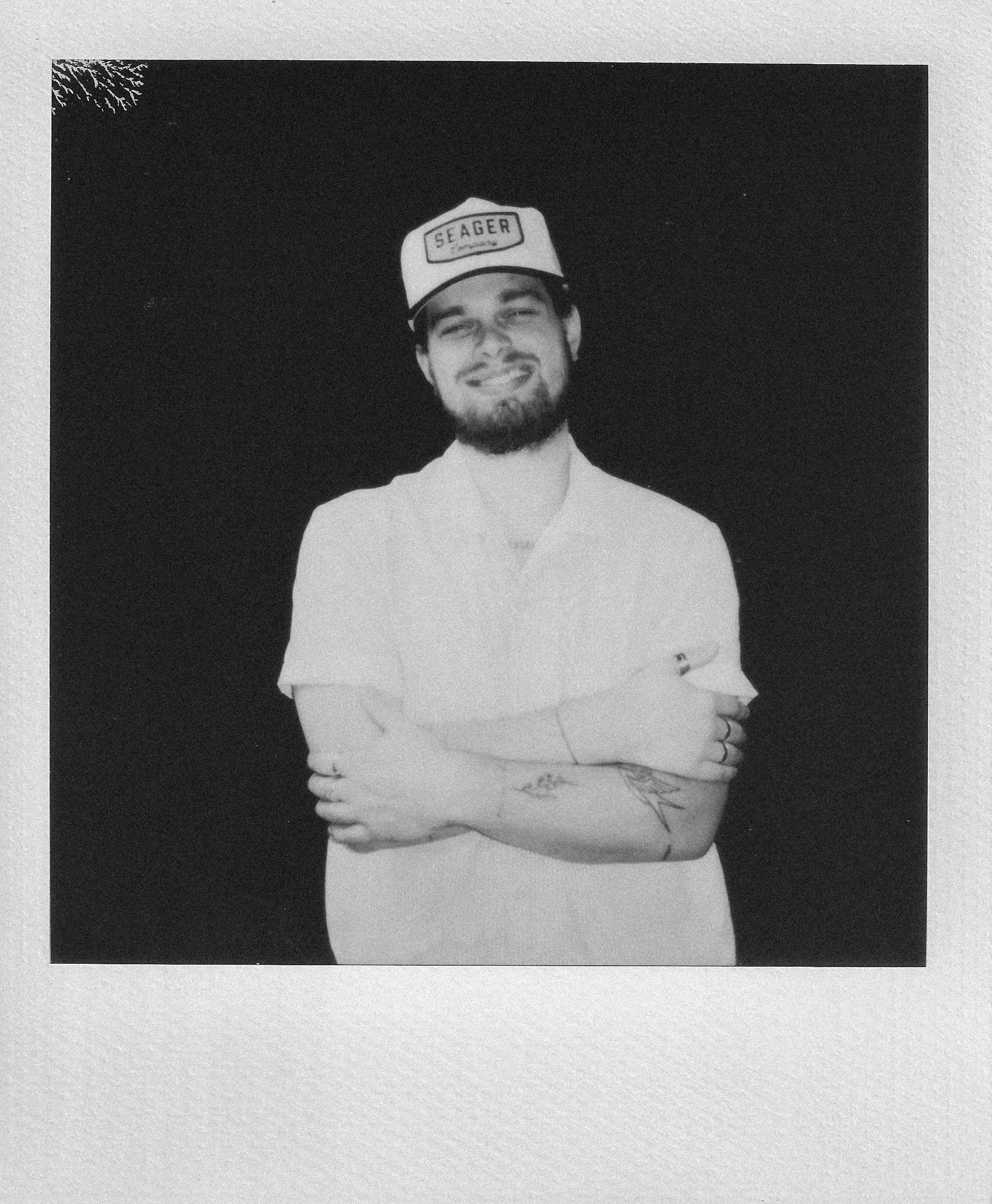 A young man with a beard and tattoos on his arms, smiling with arms crossed, wearing a cap that says "Seager," and a light-colored shirt, standing against a plain dark background.