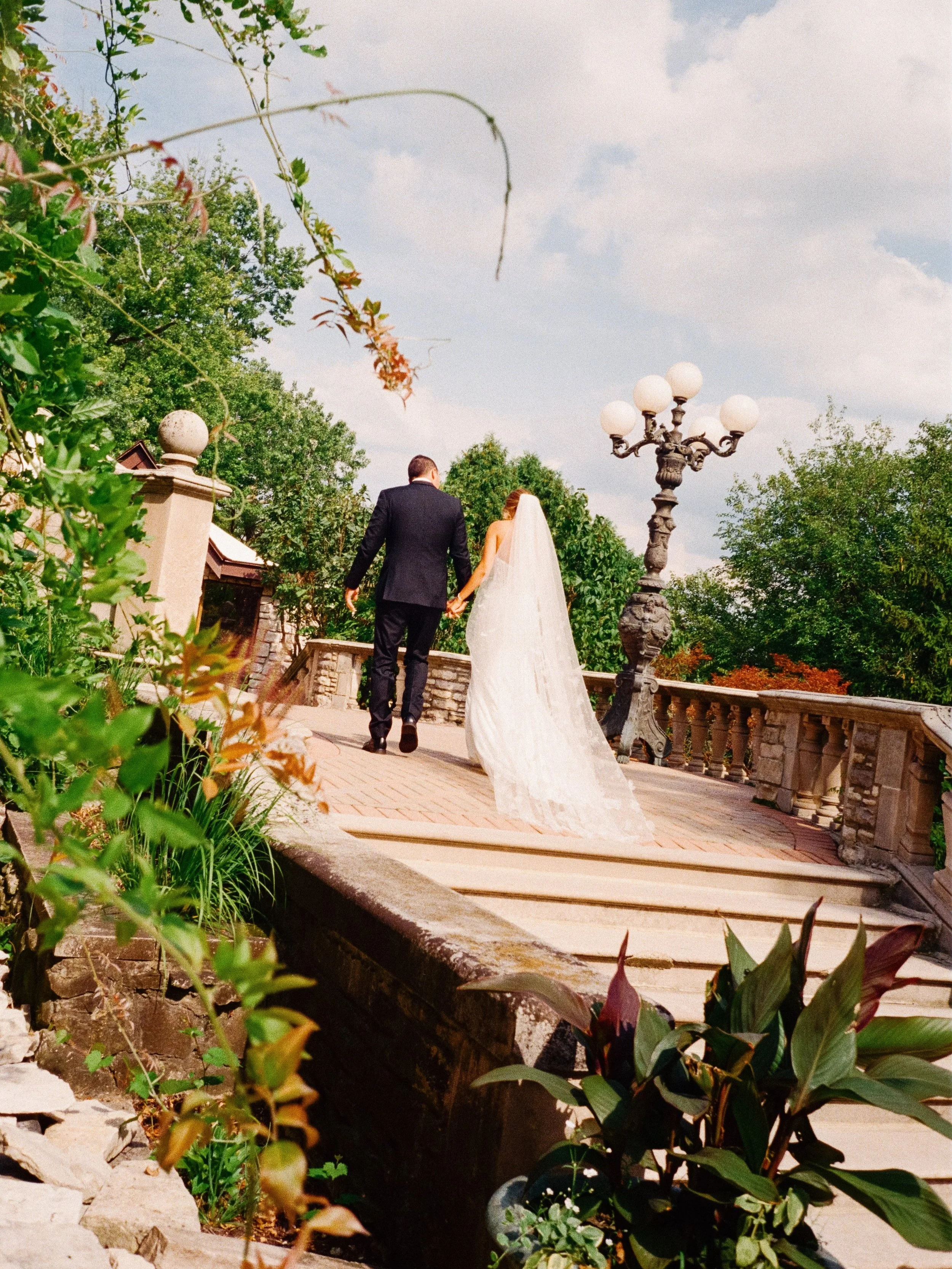 Bride and groom holding hands walking away on a brick pathway, with lush greenery and a decorative streetlamp on a cloudy day.
