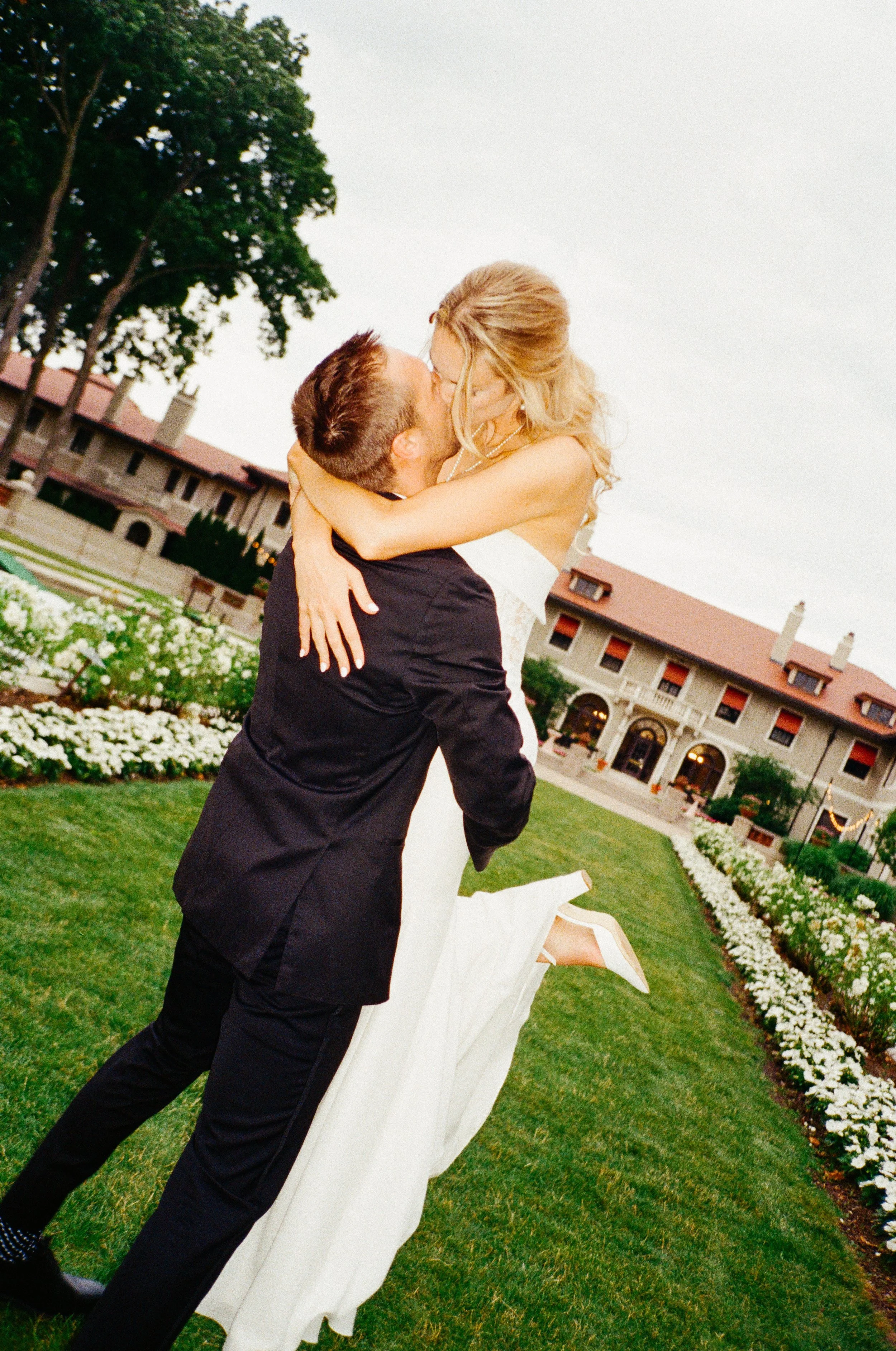 A groom lifts and kisses a bride on her wedding day outdoors in front of a large building and flower beds.
