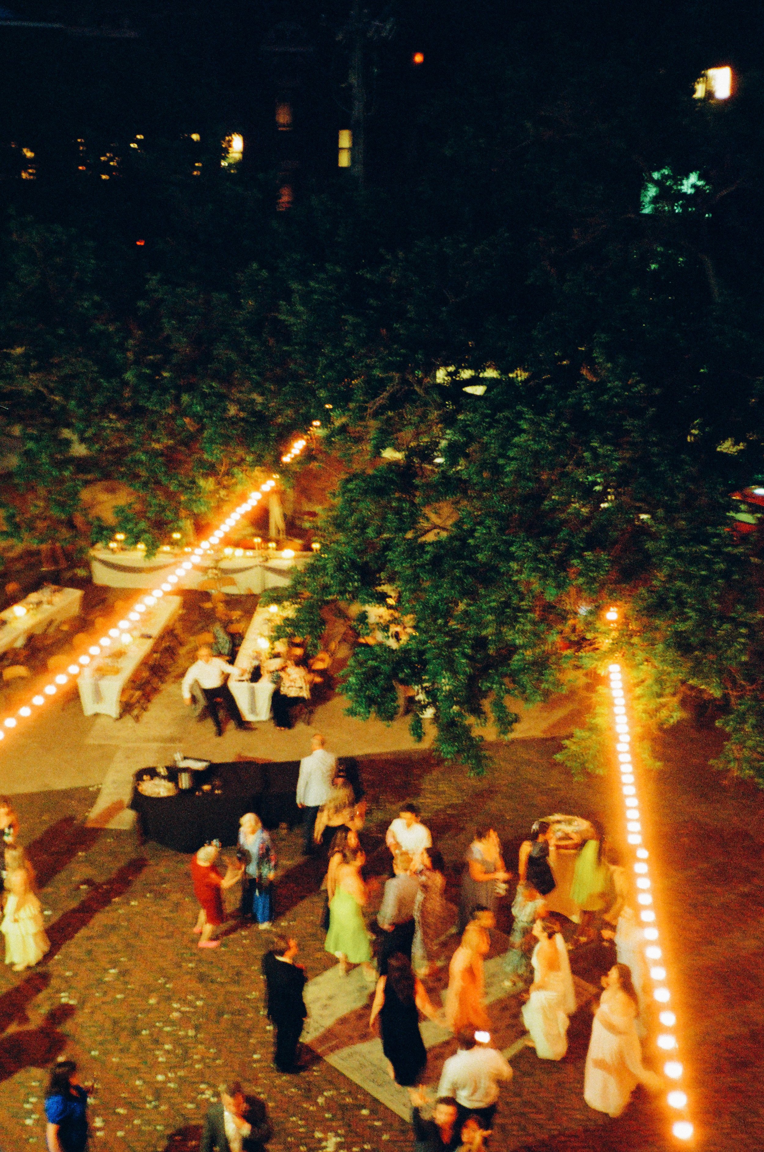 People dancing on a brick patio under string lights at an outdoor evening wedding reception with a large tree in the background.