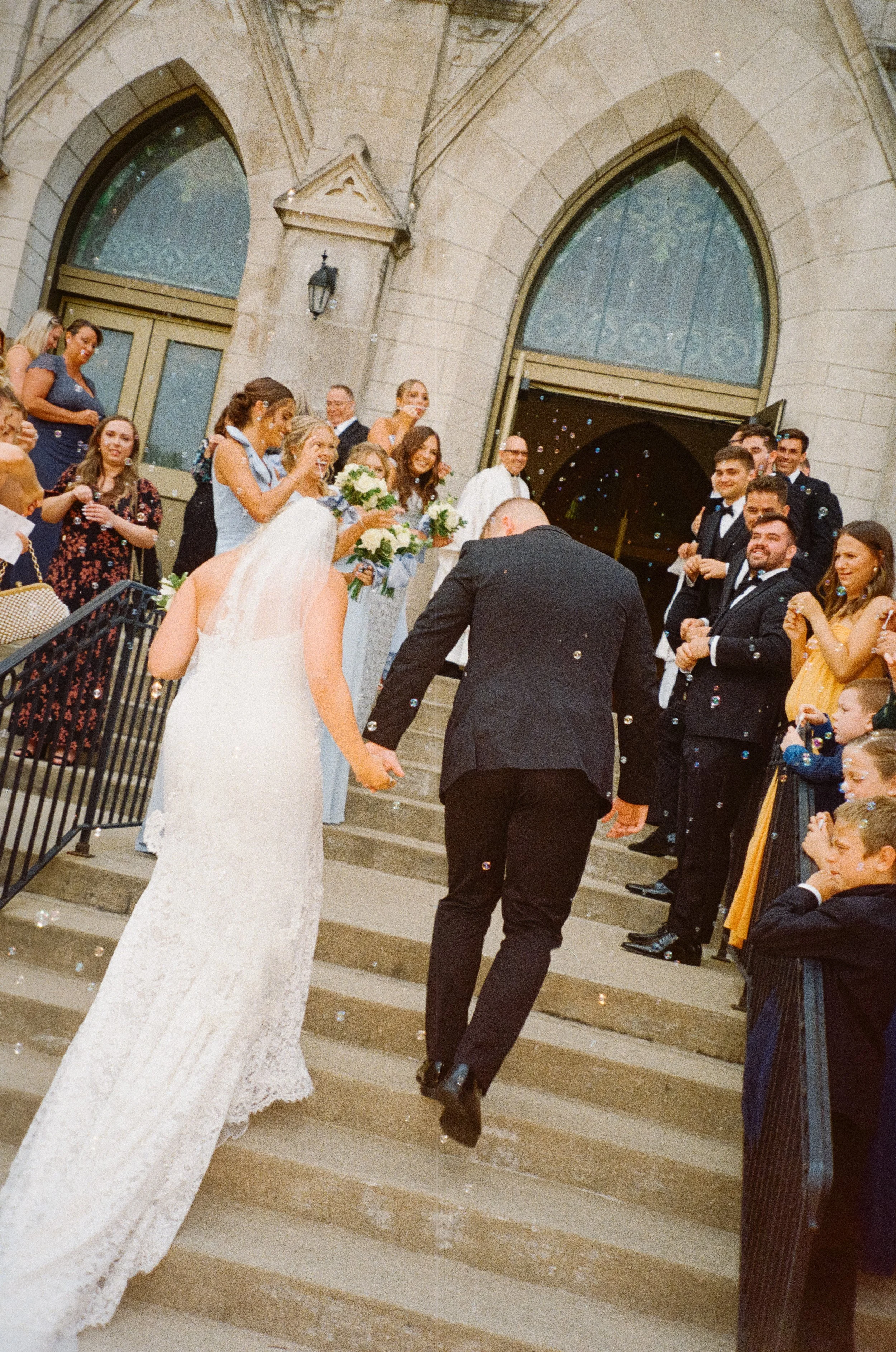 Bride and groom walking up church stairs holding hands, surrounded by wedding guests celebrating and blowing bubbles