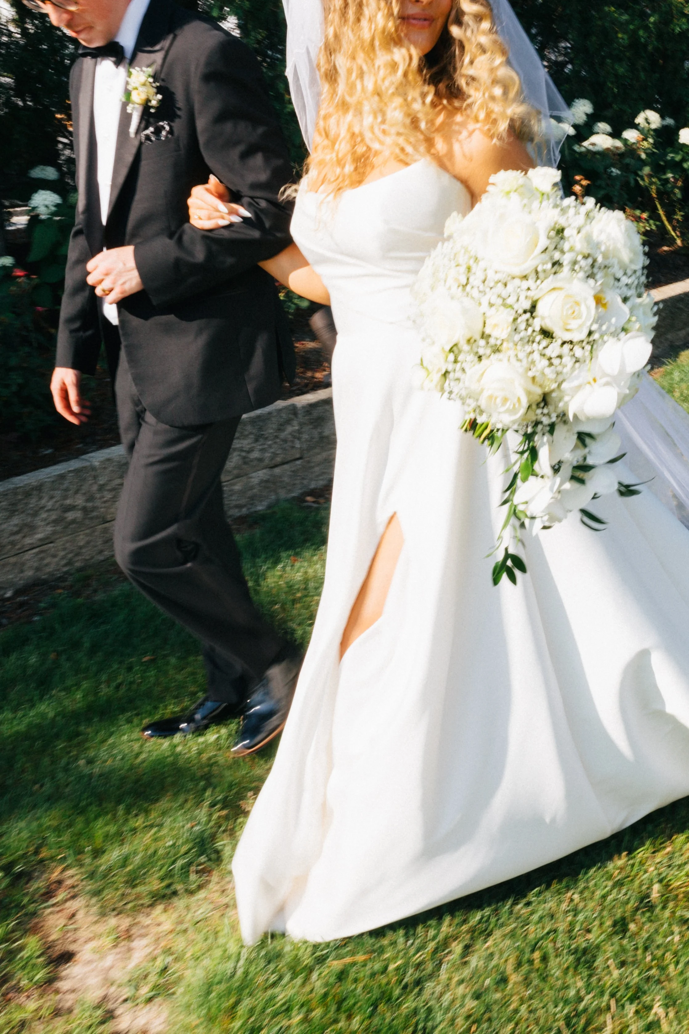 A bride and groom walking outdoors, with the bride holding a large bouquet of white flowers and wearing a white wedding gown, while the groom is dressed in a black tuxedo.