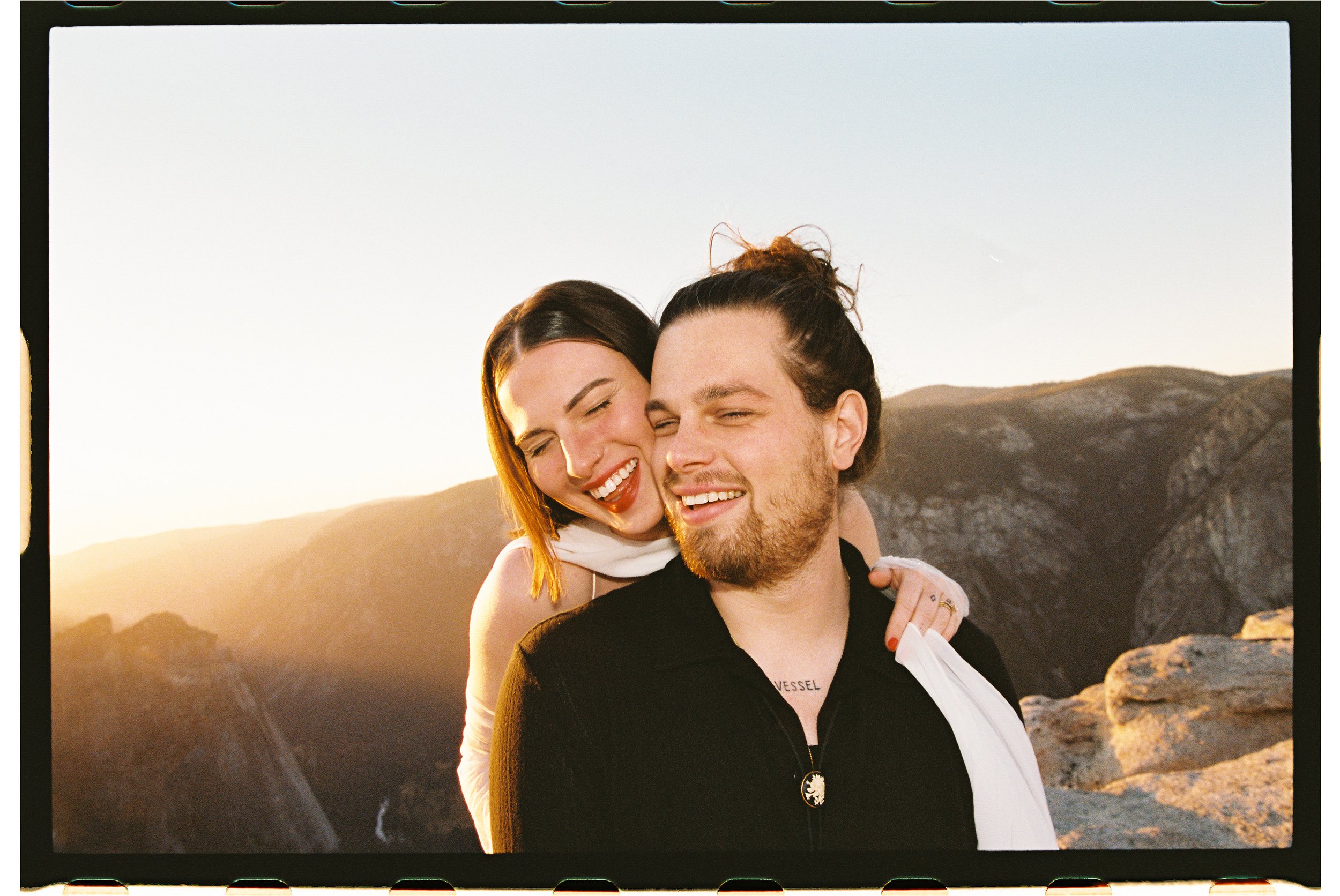 A smiling couple embracing outdoors at sunset, with mountains and a canyon in the background.