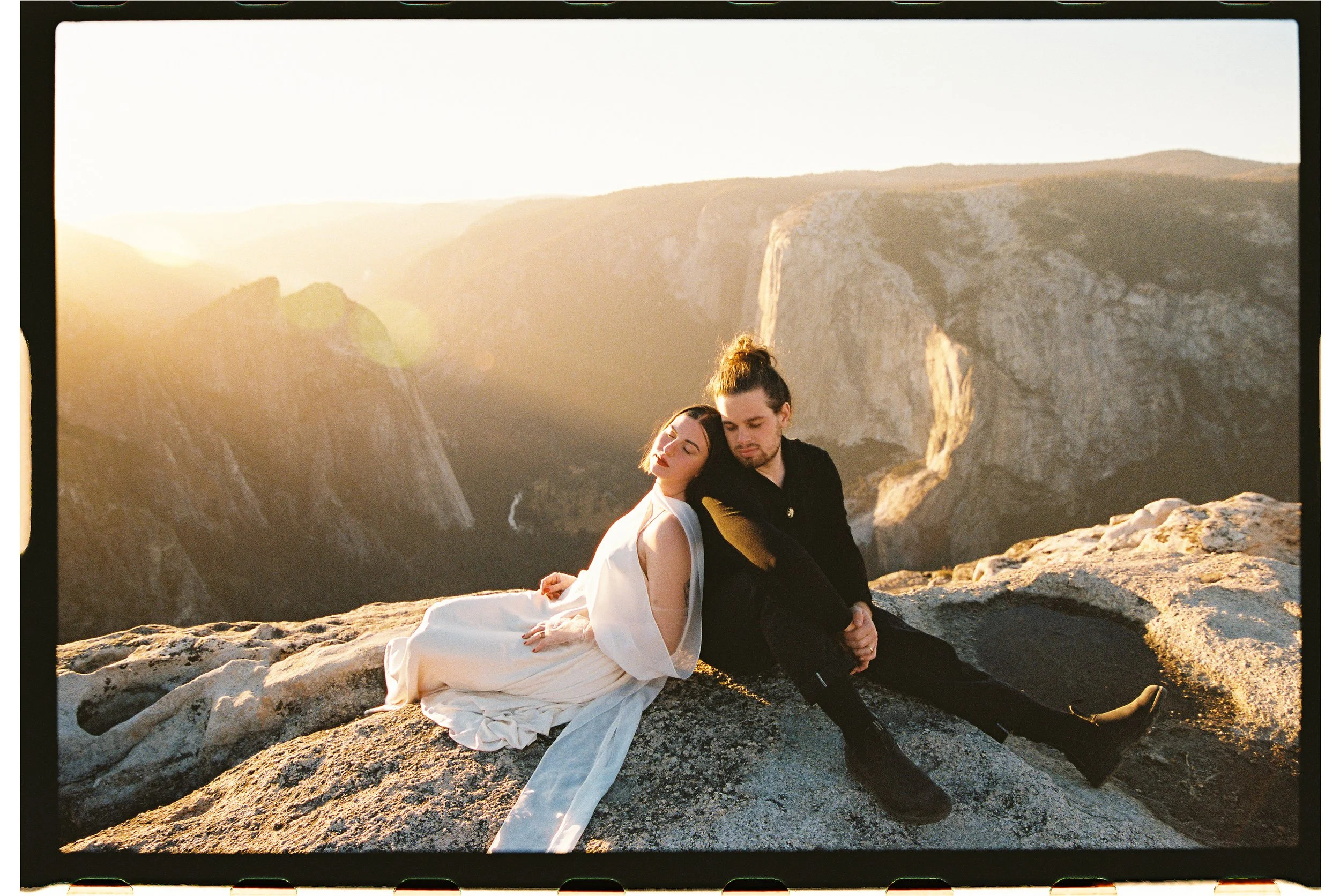 A man and woman sitting on a rock at sunset in a mountainous landscape, with the woman leaning on the man's shoulder.
