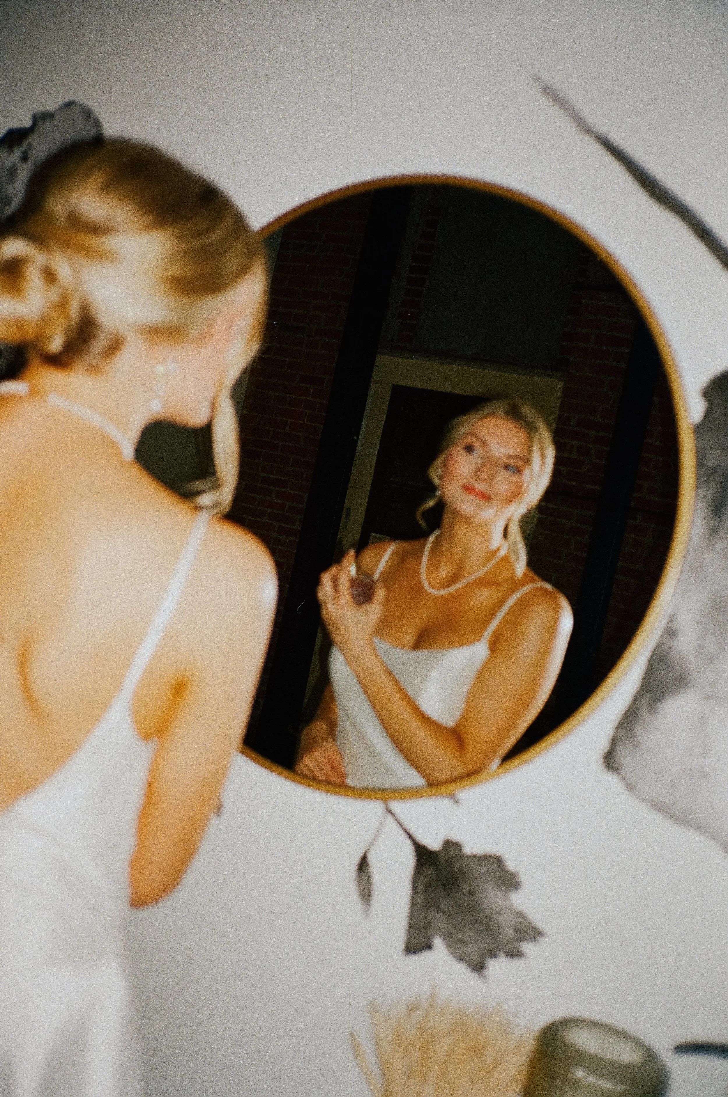 A woman in a white dress and pearl jewelry looks at her reflection in a round mirror, holding a makeup brush in her right hand.