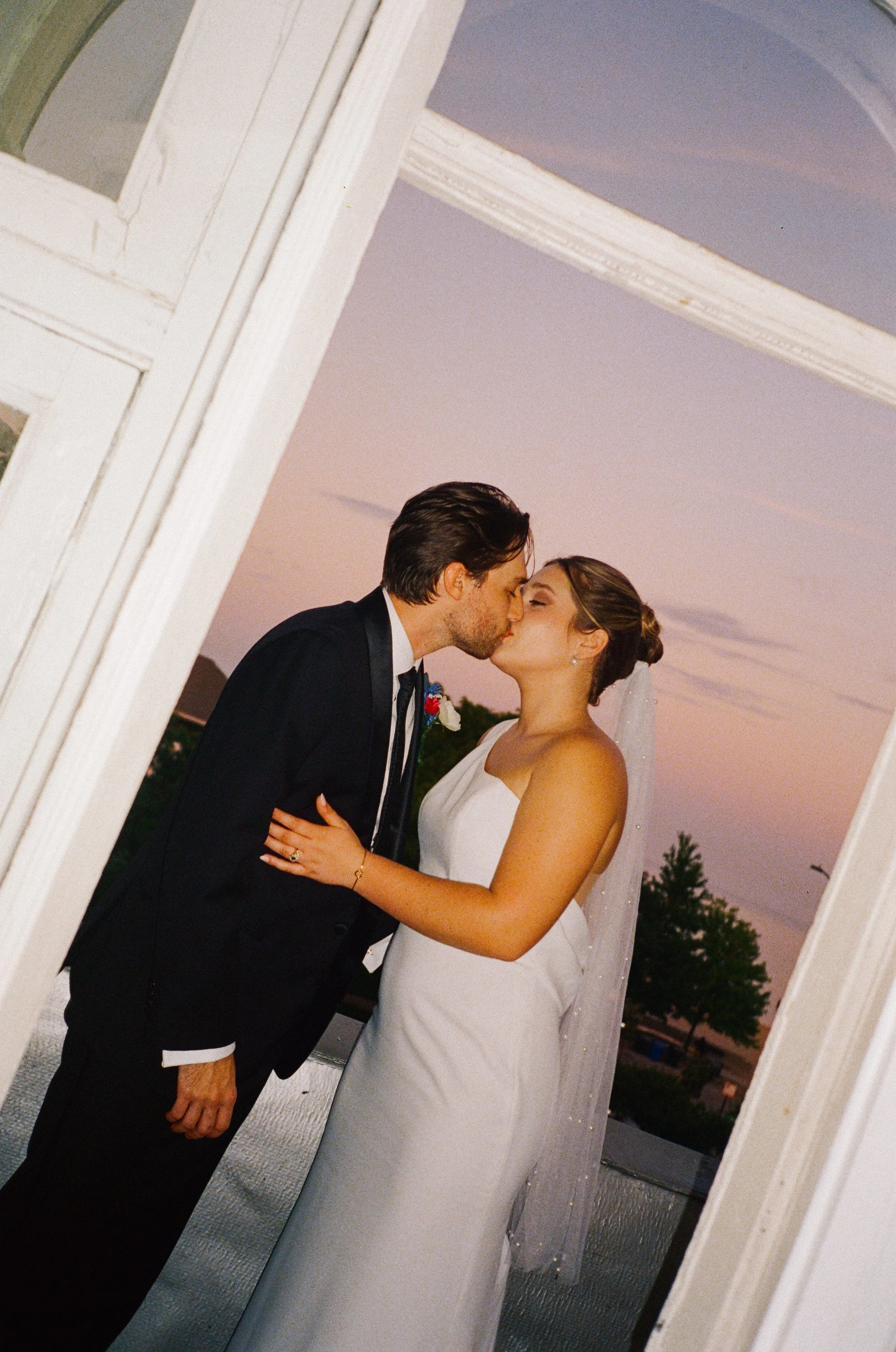 A couple in wedding attire sharing a kiss on a balcony at sunset, framed by a window.