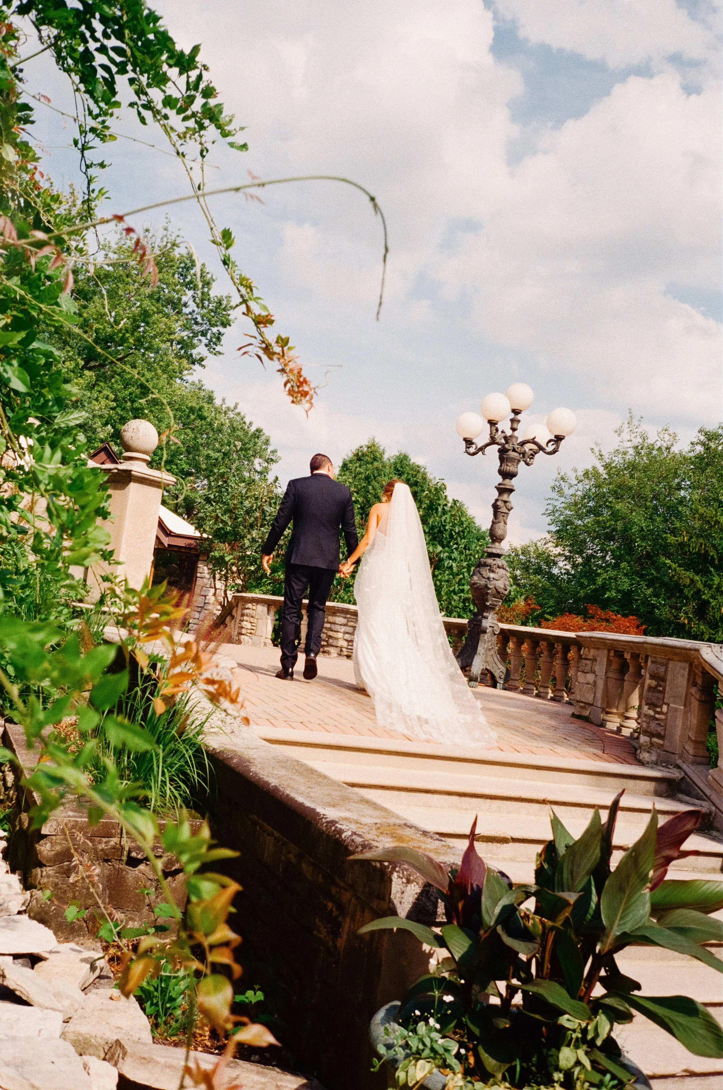 A bride and groom holding hands as they walk away on a stone bridge surrounded by greenery and trees, with a cloudy sky overhead.