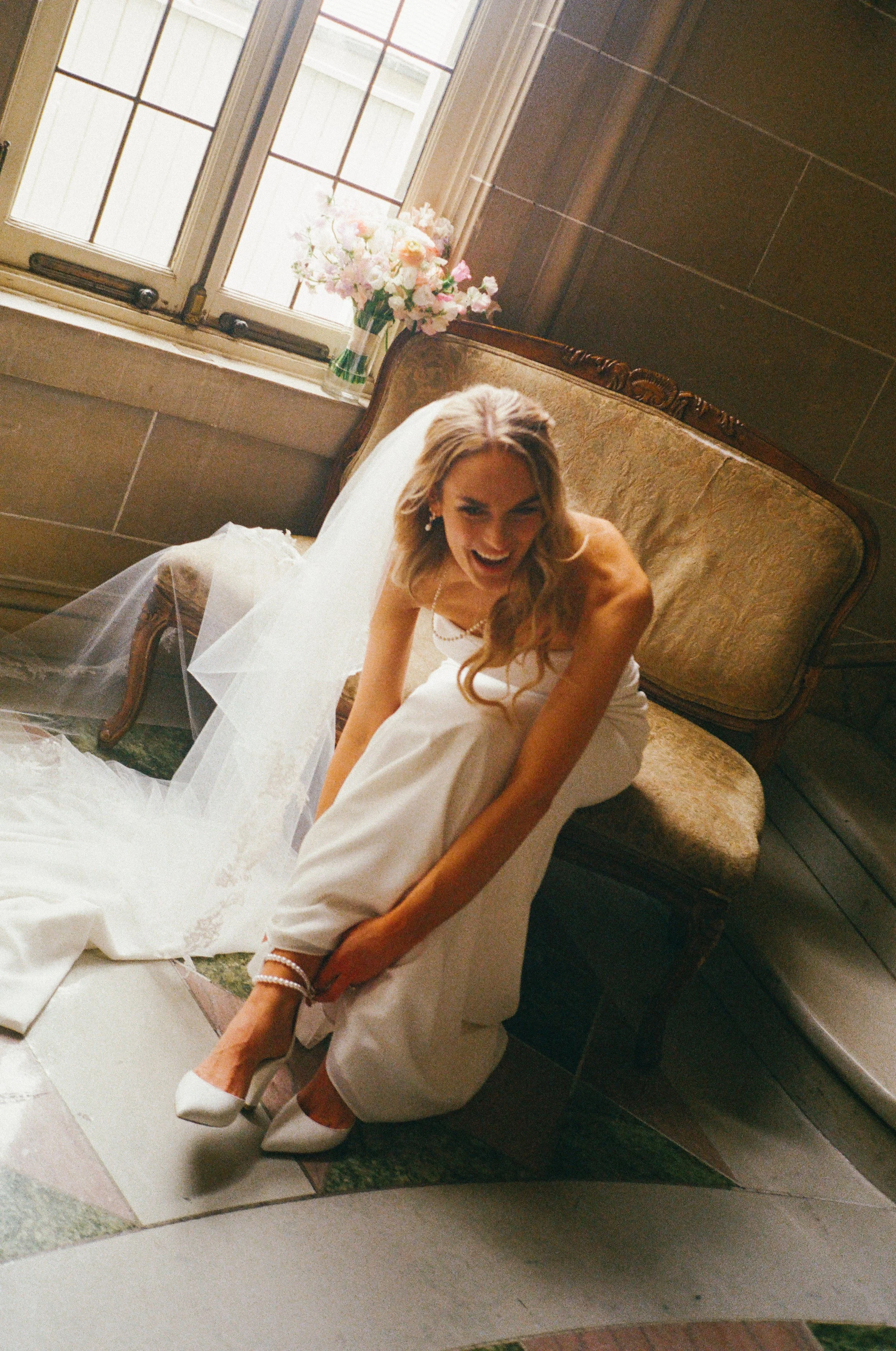 A smiling bride in a white wedding dress sits on a vintage sofa near a window with a flower bouquet nearby.