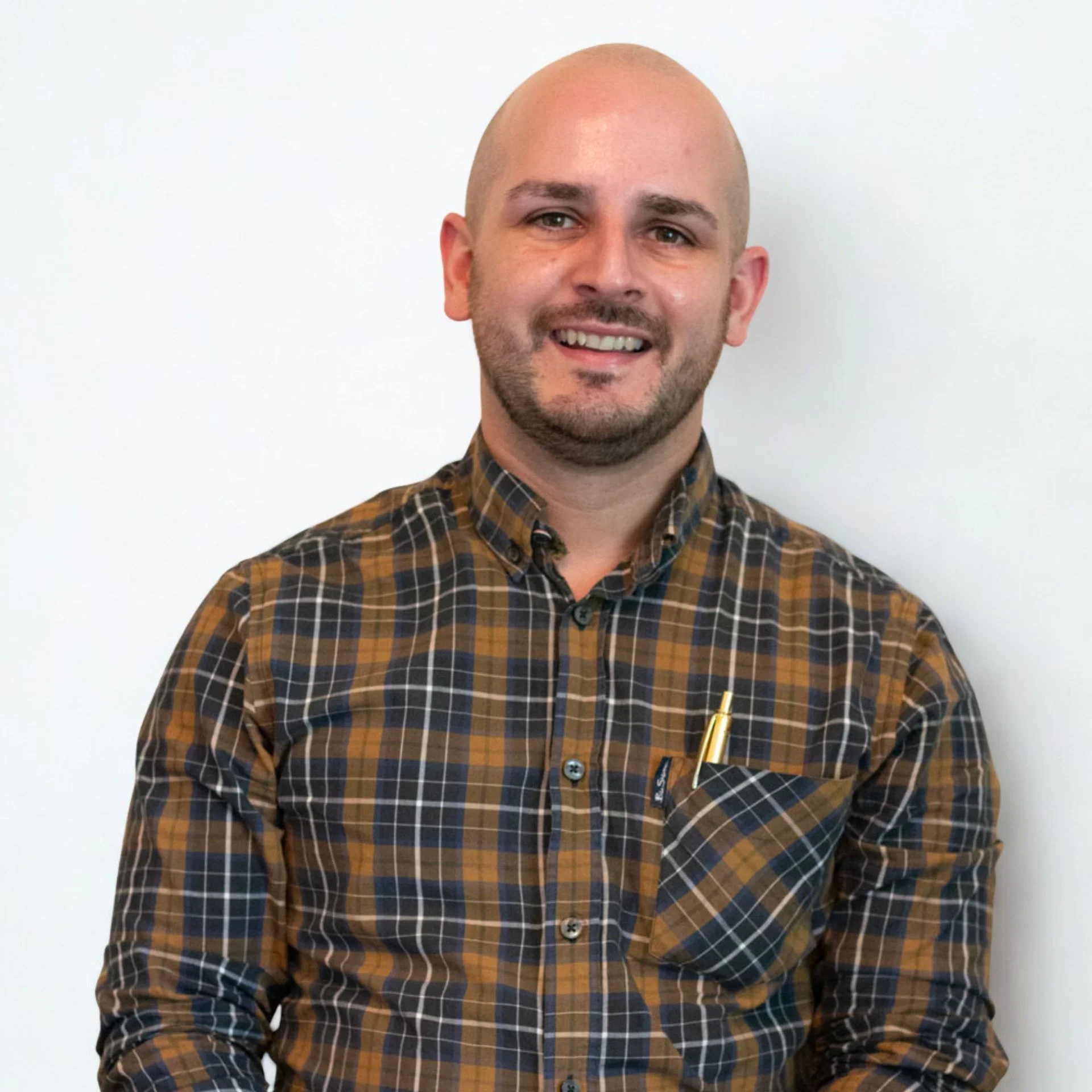 A man with a shaved head, beard, wearing a brown plaid shirt with a pen in the chest pocket, smiling against a plain white wall.
