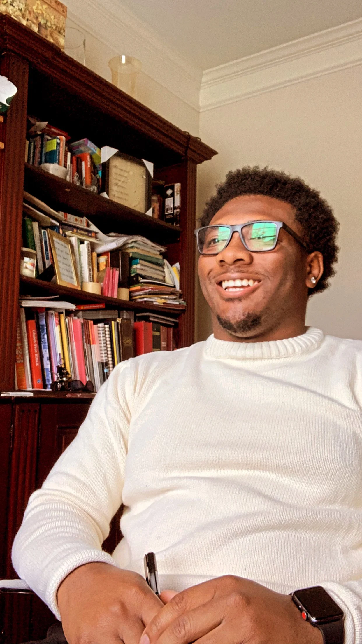 A smiling man with glasses and a white sweater sitting in front of a wooden bookshelf filled with books and papers.