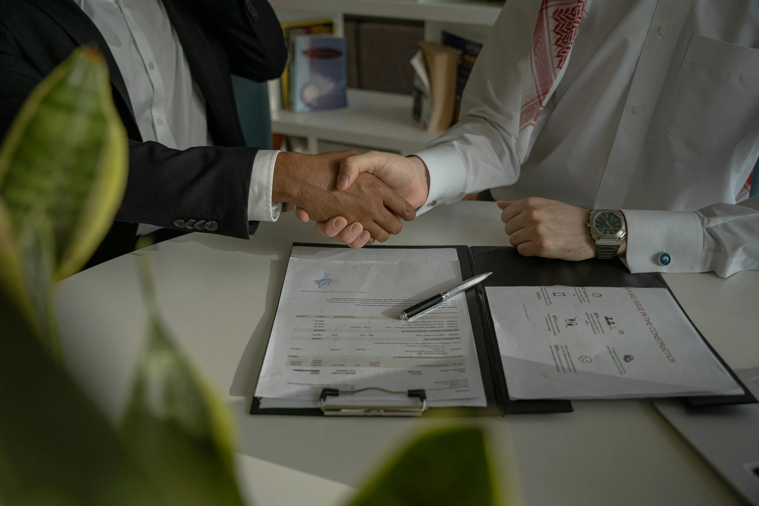 Two men in business attire shaking hands across a desk, with documents, a pen, and a clipboard on the table, in an office setting.