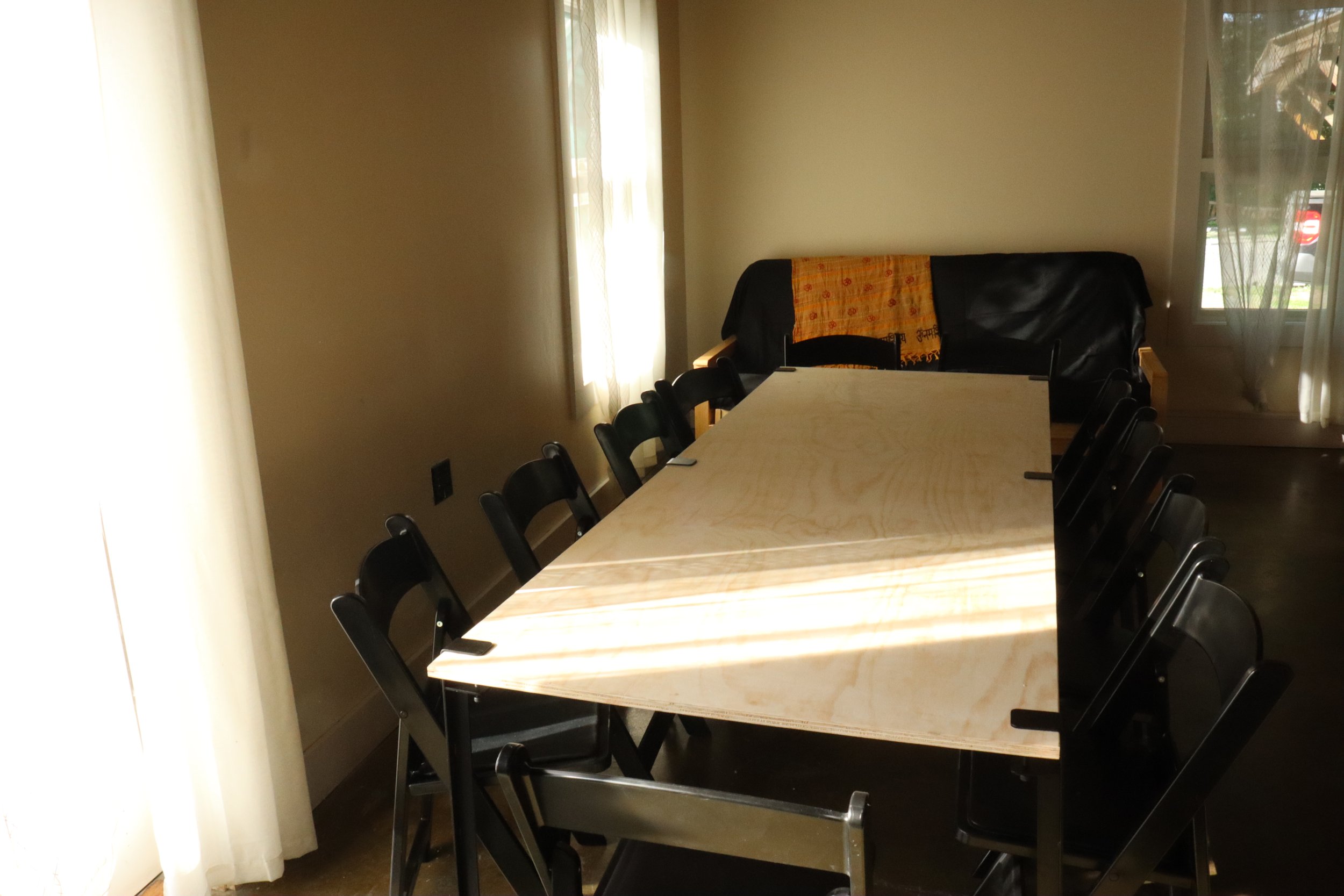 A conference room with a long wooden table surrounded by black chairs. There is a black couch against the wall with an orange and yellow cloth draped over the back. Two large windows with sheer white curtains let in natural light.
