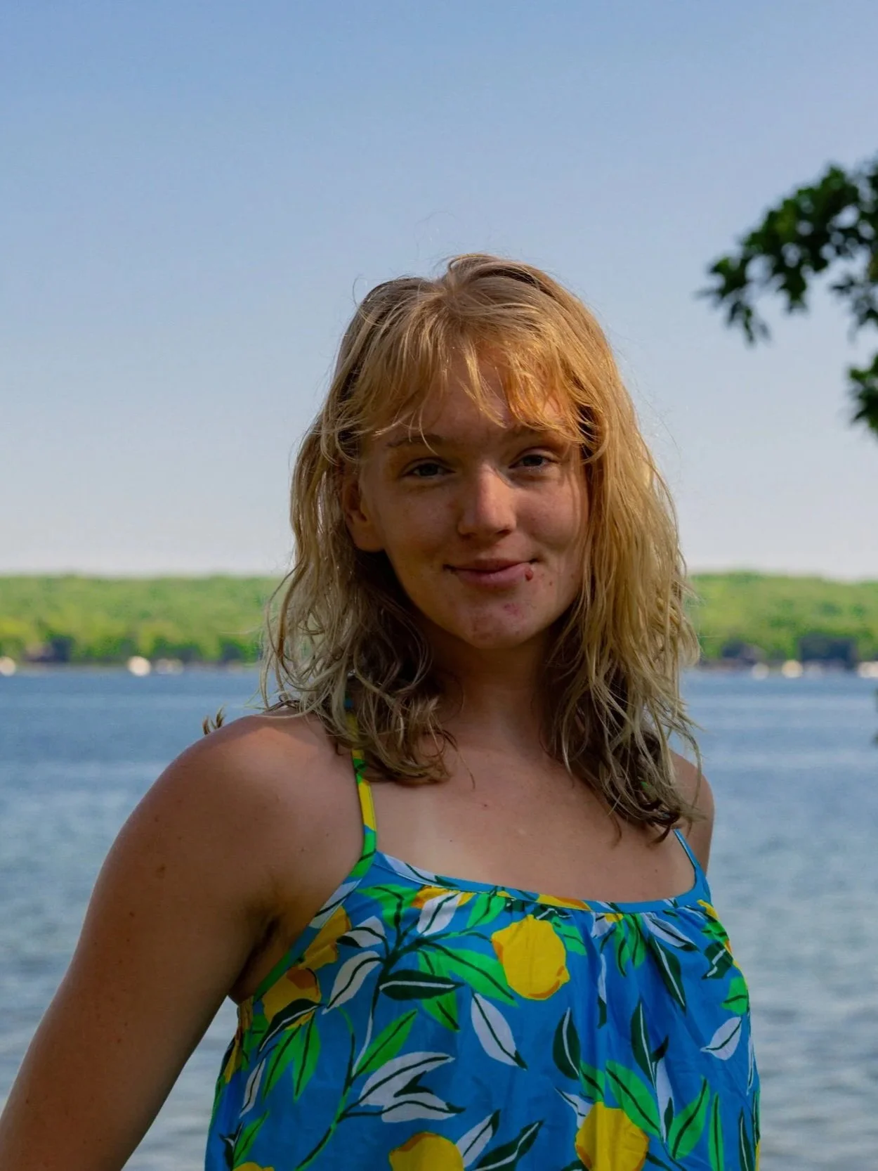 A young woman with shoulder-length blonde hair standing by a lake, wearing a colorful tank top with a lemon and leaf pattern, smiling with a background of calm water and lush green trees.