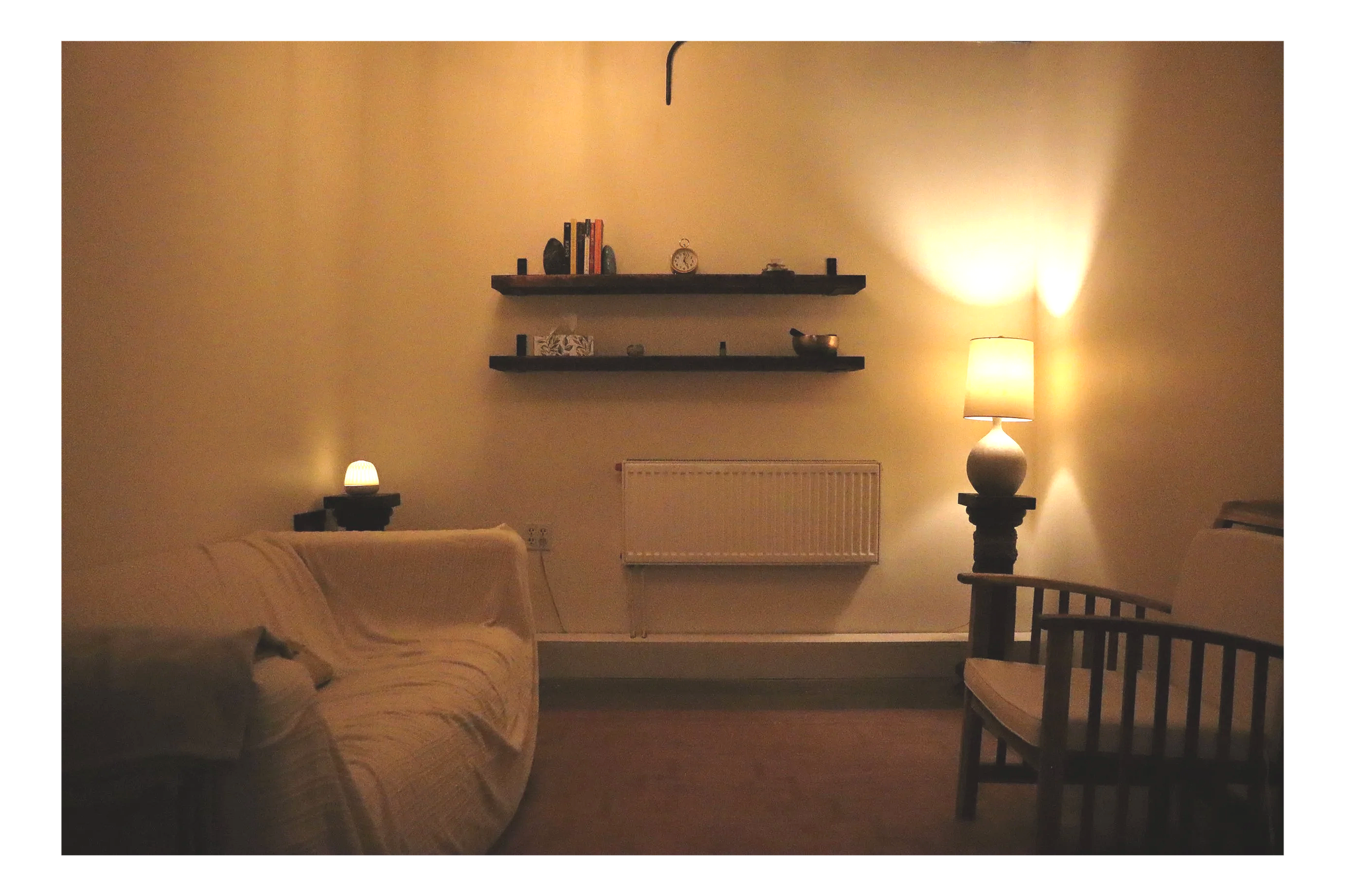 A dimly lit living room with two floating shelves on a yellowish wall, a clock, books, and decorative items, a standing lamp emitting warm light, a small lamp on a side table, a white radiator, a beige couch, and a wooden chair.