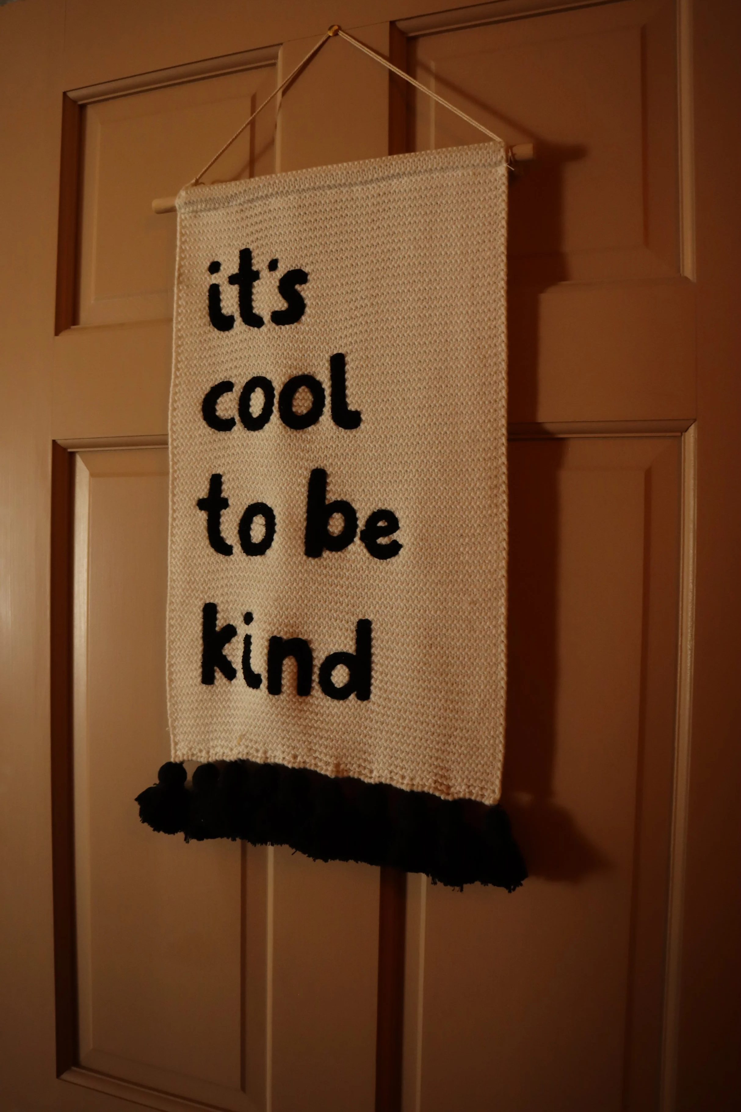 A fabric banner hanging on a door with the words 'it's cool to be kind' embroidered in black letters. The banner has black pom-poms at the bottom.