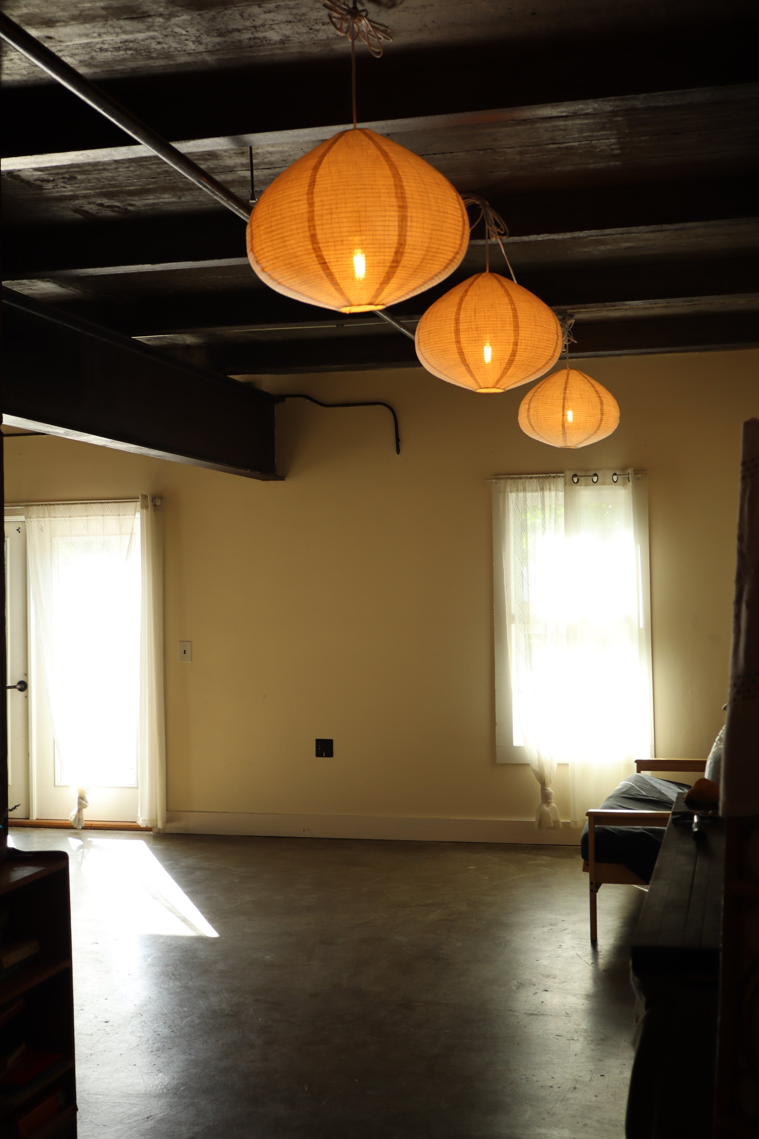 A room with a dark wooden ceiling, beige walls, and large windows covered with sheer curtains. Three hanging paper lanterns emit warm light. There is a couch on the right side and some furniture, with sunlight streaming through the windows.