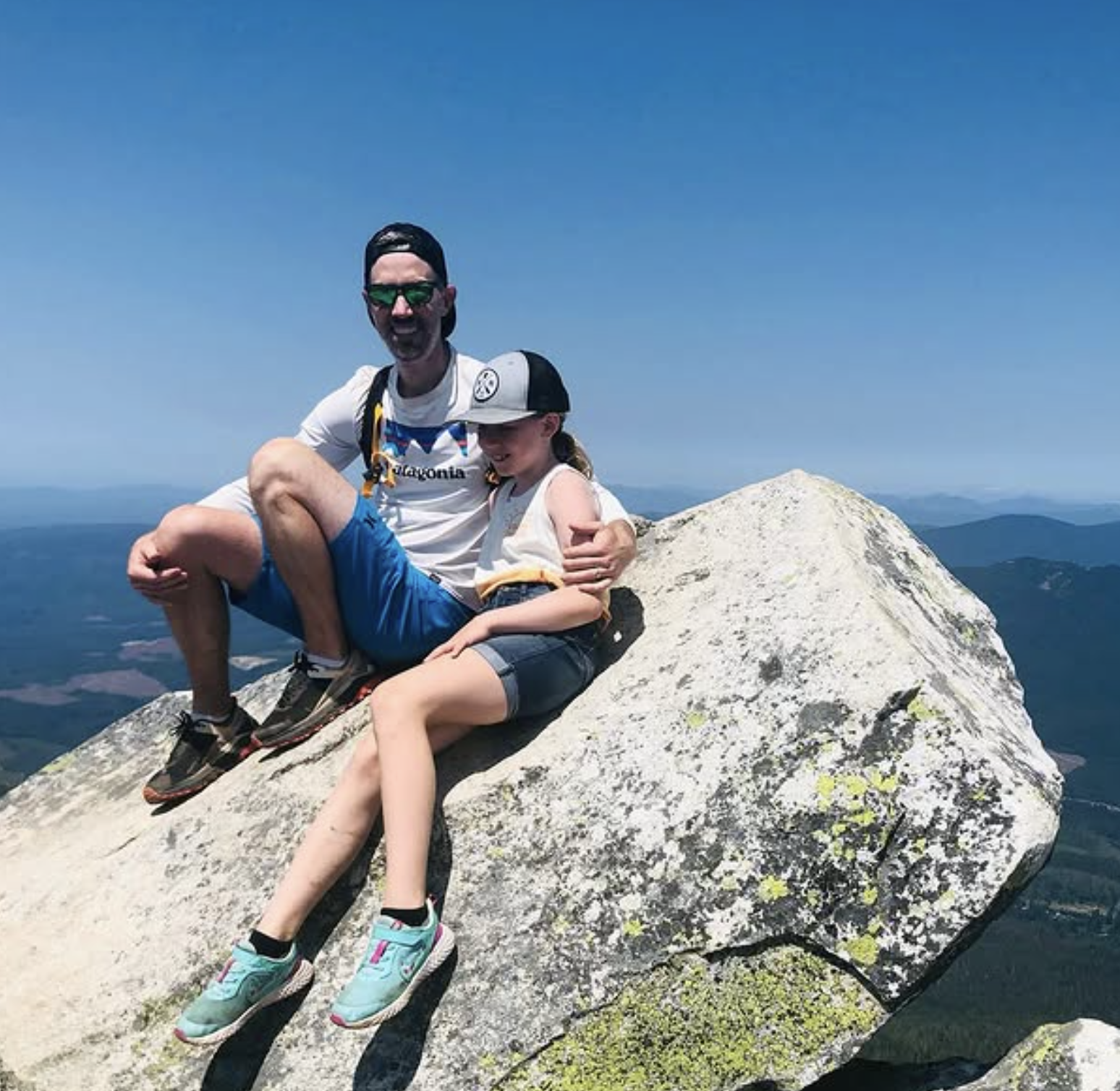 A man and a young girl sitting on a large white rock at the top of a mountain, smiling with mountains and blue sky in the background.