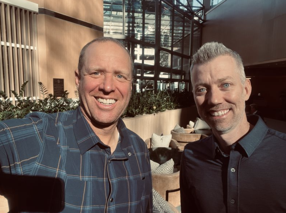 Two smiling men taking a selfie in a modern, sunny indoor setting with large glass windows, plants, and seating in the background.