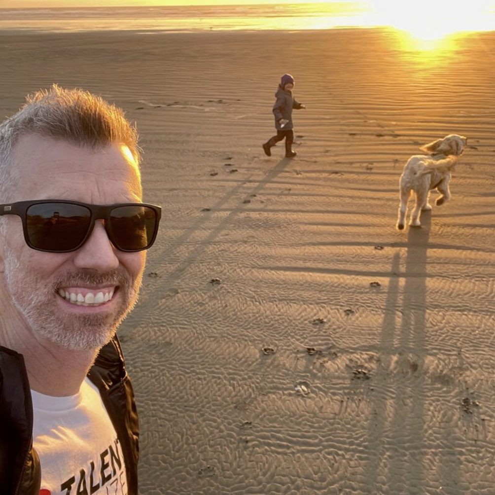 A man wearing sunglasses taking a selfie on a sandy beach at sunset, with two people walking dogs in the background.