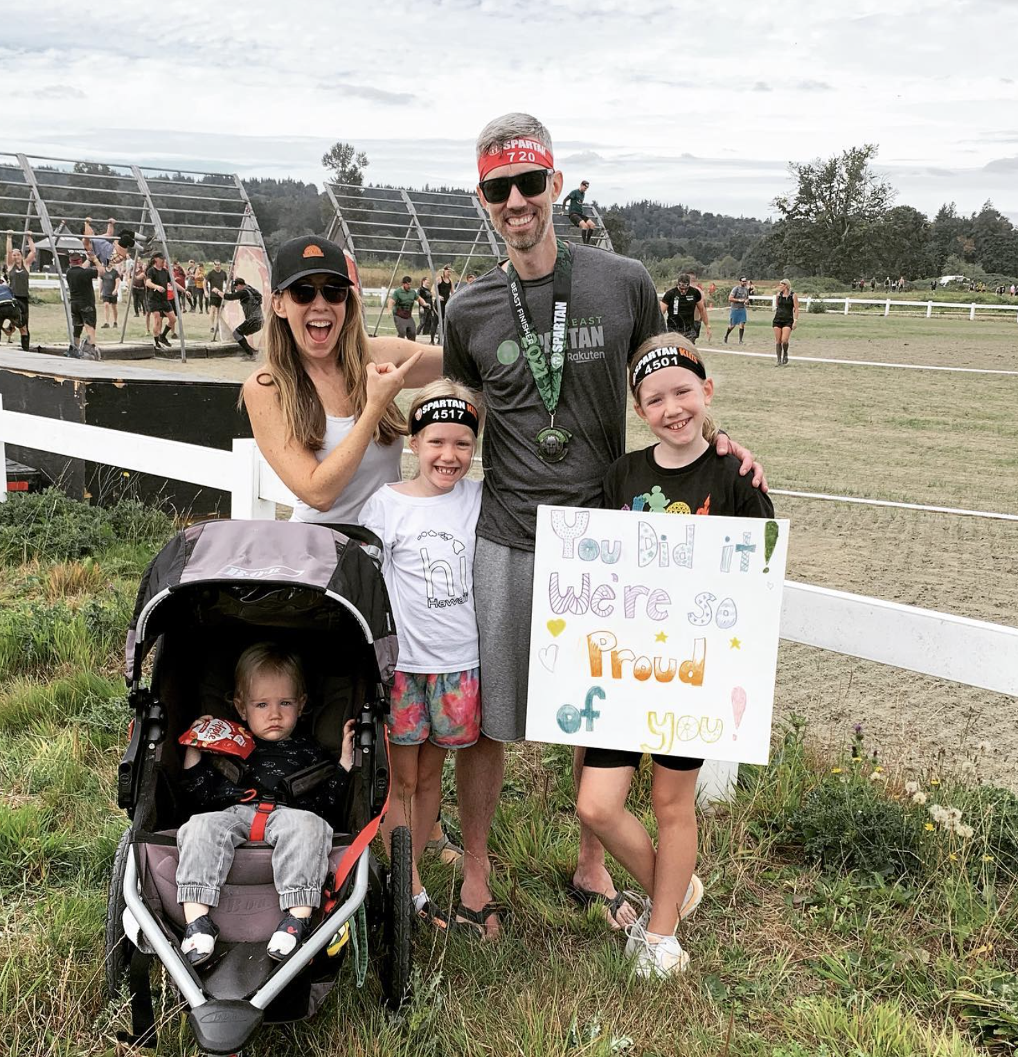 A family celebrating after a running event, with a man wearing a finisher's medal and children holding a proud sign, with others participating in a race in the background.