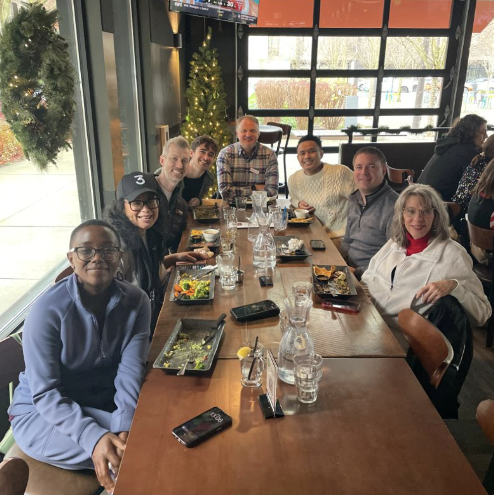 A group of nine people sitting around a long wooden table in a restaurant, celebrating with food and drinks. In the background, holiday decorations including a Christmas tree and a wreath are visible.