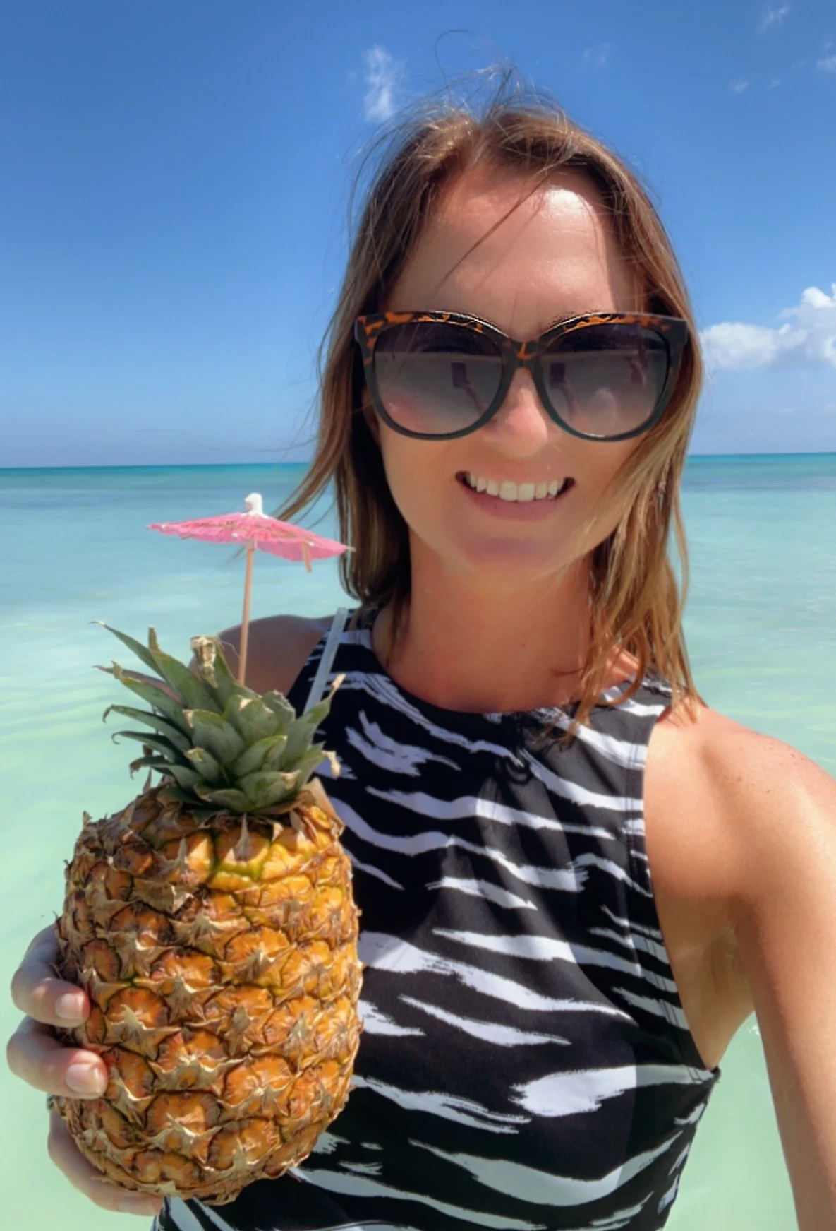 Woman with sunglasses smiling at the beach, holding a pineapple with a small pink umbrella in it.
