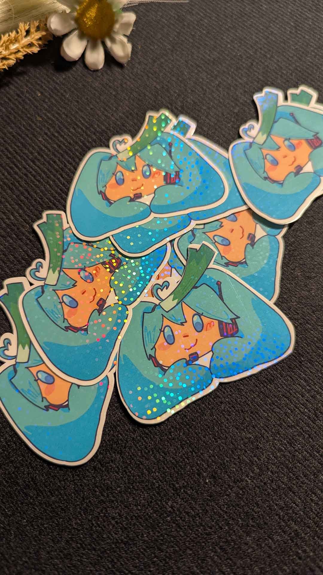 A pile of holographic stickers featuring a cartoon character named Hatsume Miku, with blue hair, blue eyes, and a cheerful expression, on a black surface with a small white flower and dried plant in the background.