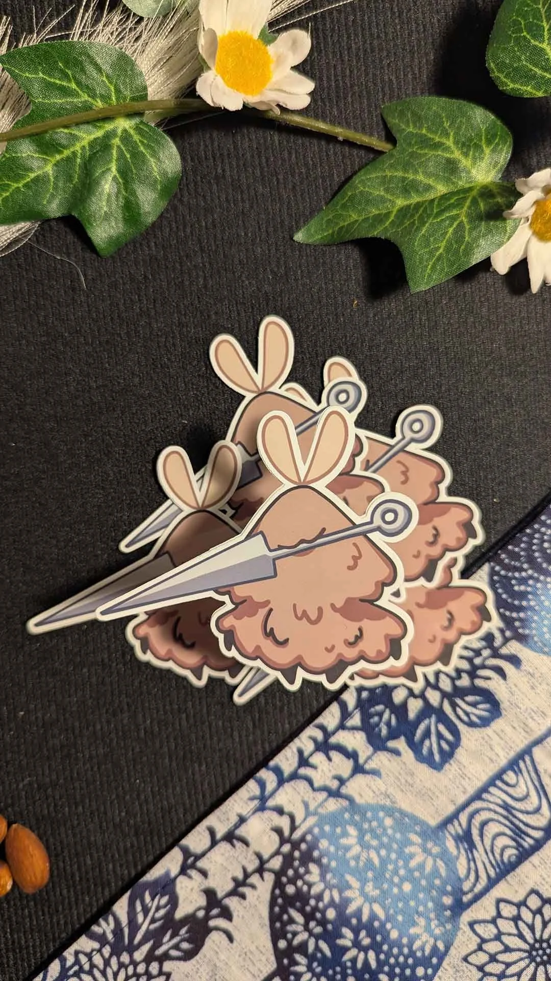 A group of stickers featuring fluffy, warm-brown fleas with long antennae, holding needles, placed on a black surface surrounded by artificial green leaves and white daisies, with a blue and white patterned fabric partially visible.