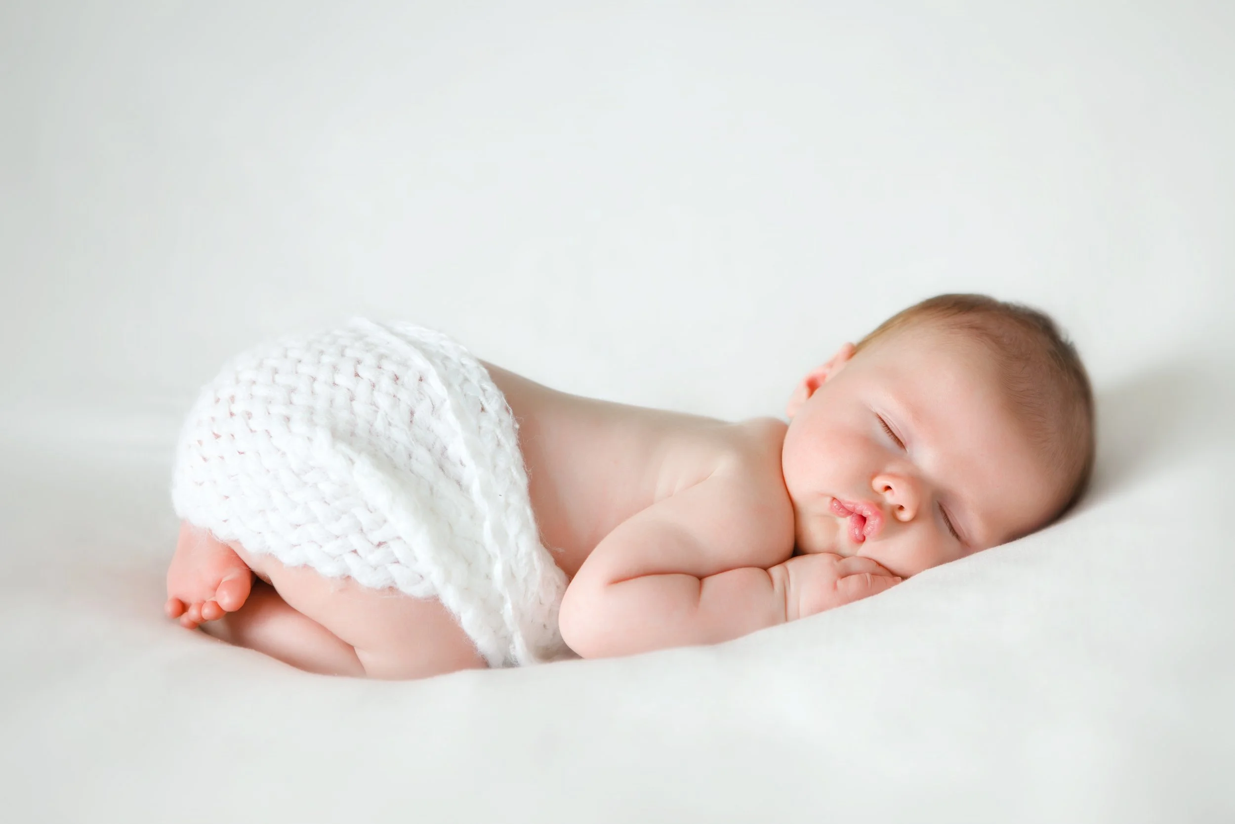 A sleeping baby lying on its side on a white surface, wearing a white knitted diaper cover, with arms tucked under its head, and a peaceful expression