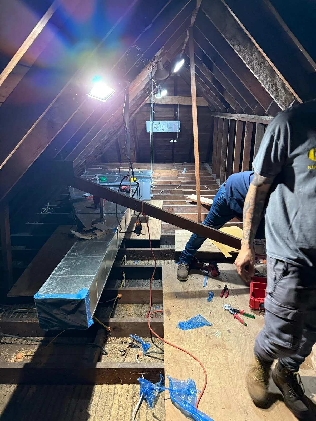 Construction workers installing ductwork in an attic space with wood framing and insulation. One worker is crouched down working on a duct, and another is partially visible on the right side.
