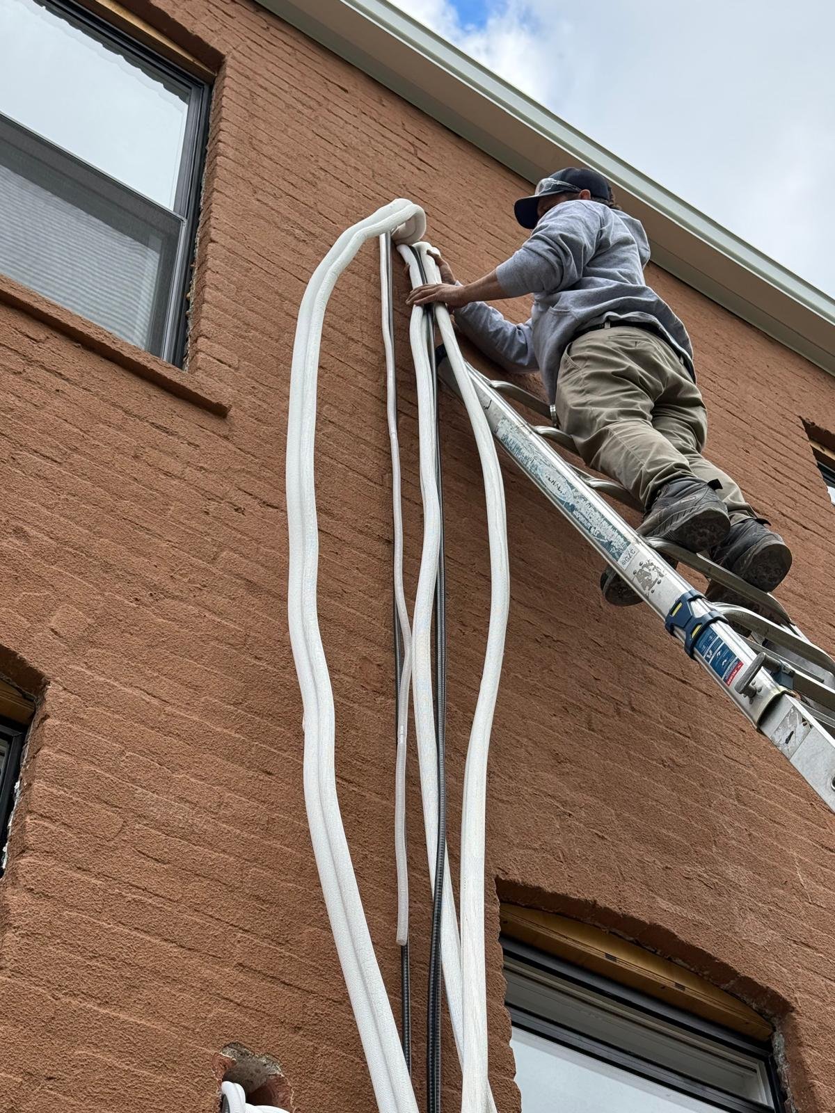 A worker on a ladder installing or repairing electrical cables on the side of a brick building.