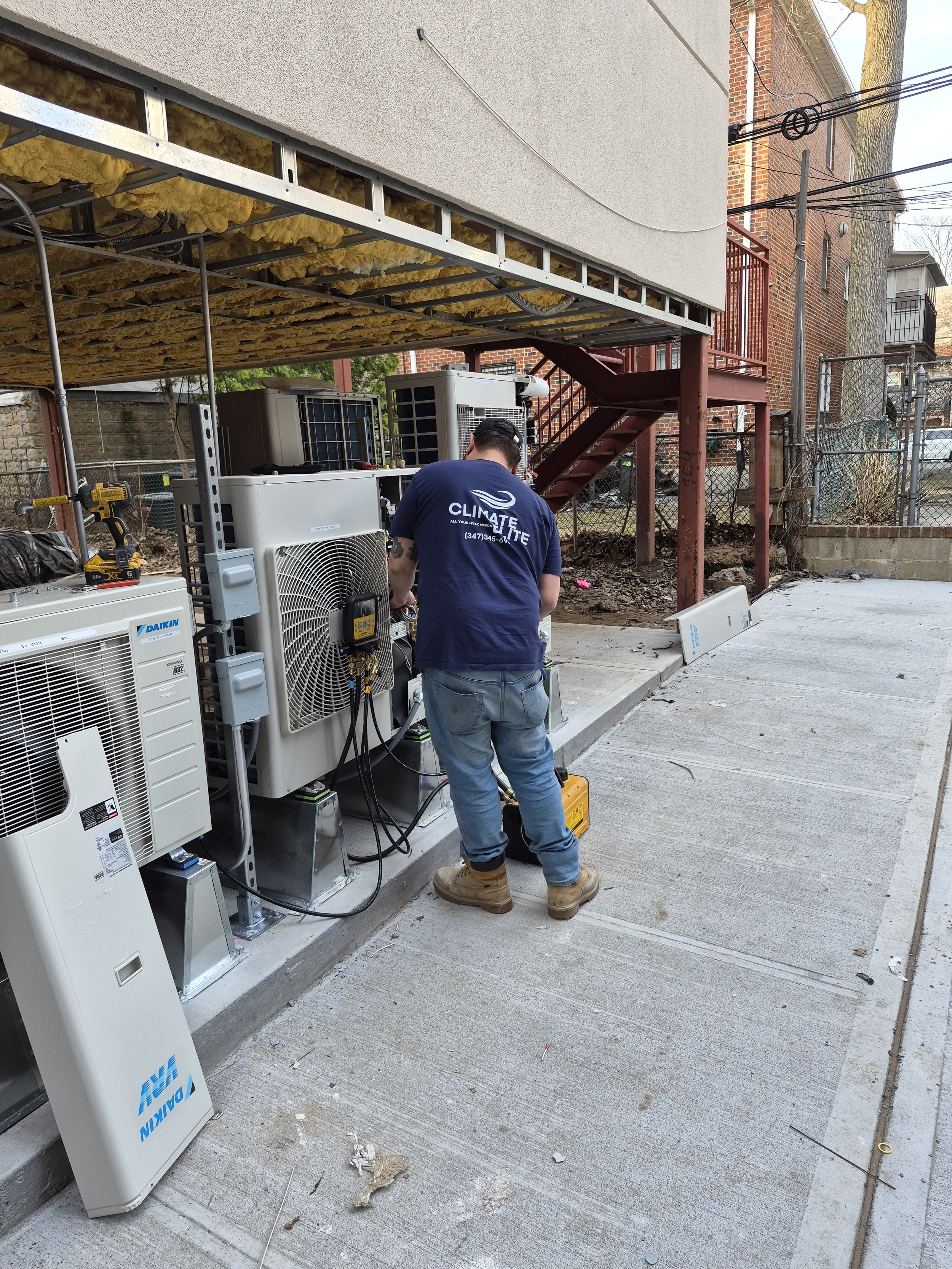 A technician working on HVAC equipment outside a building, with several air conditioning units and electrical wires visible.