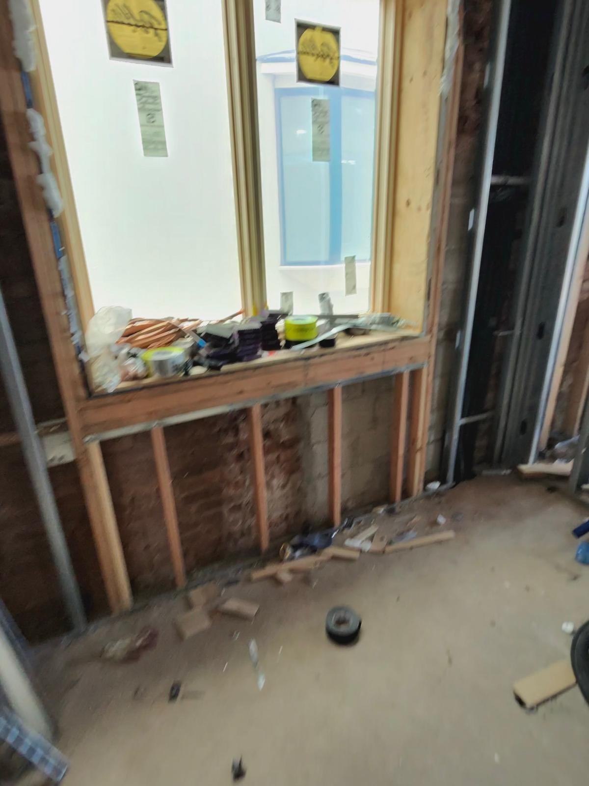 Interior of a house under construction with a window frame, construction tools, and materials on the window sill, exposed wall studs, and scattered debris on the unfinished floor.
