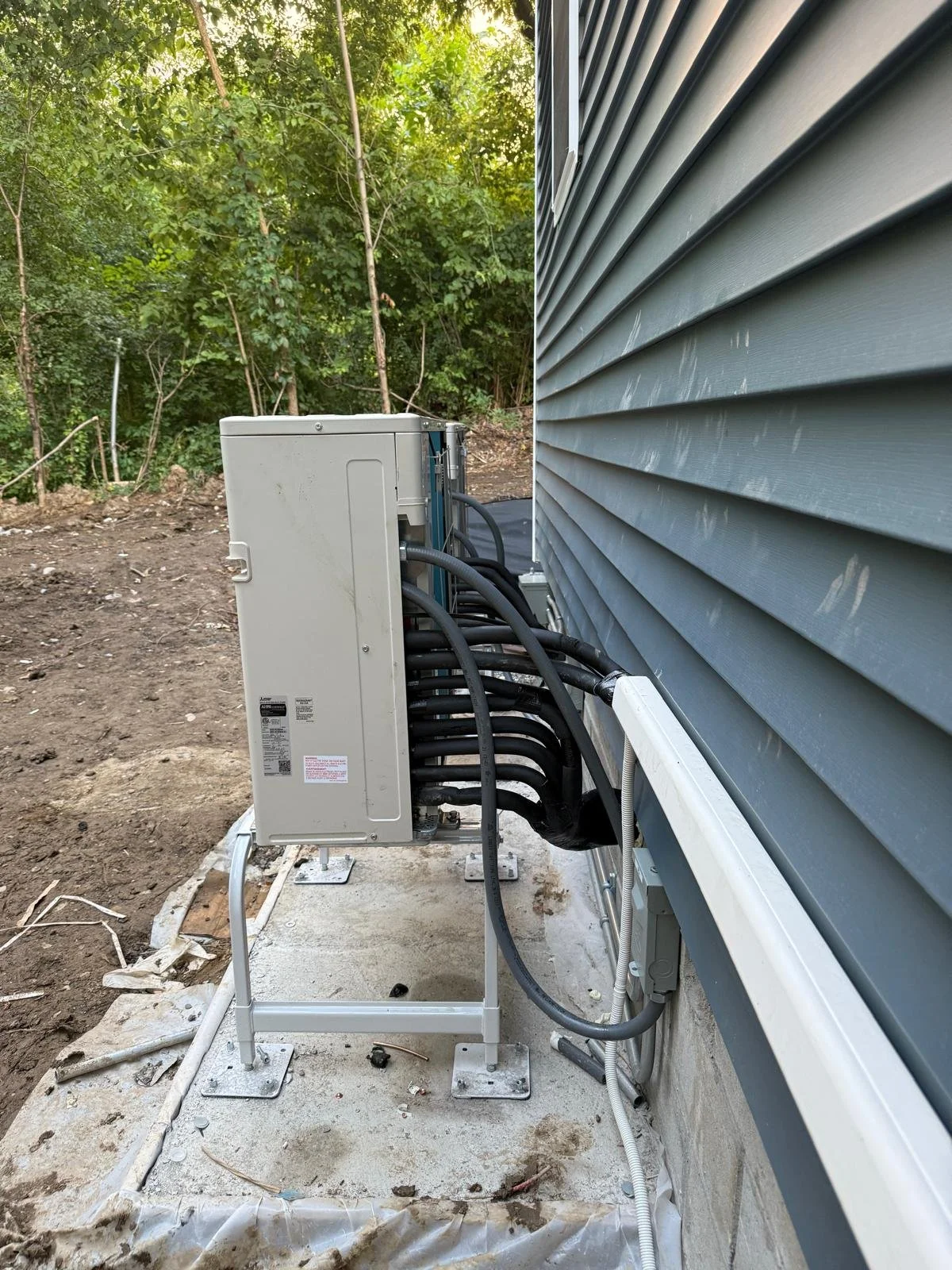 Electrical or HVAC equipment outside a building next to gray vinyl siding, with cables connected to the equipment and some dirt and debris on the ground.
