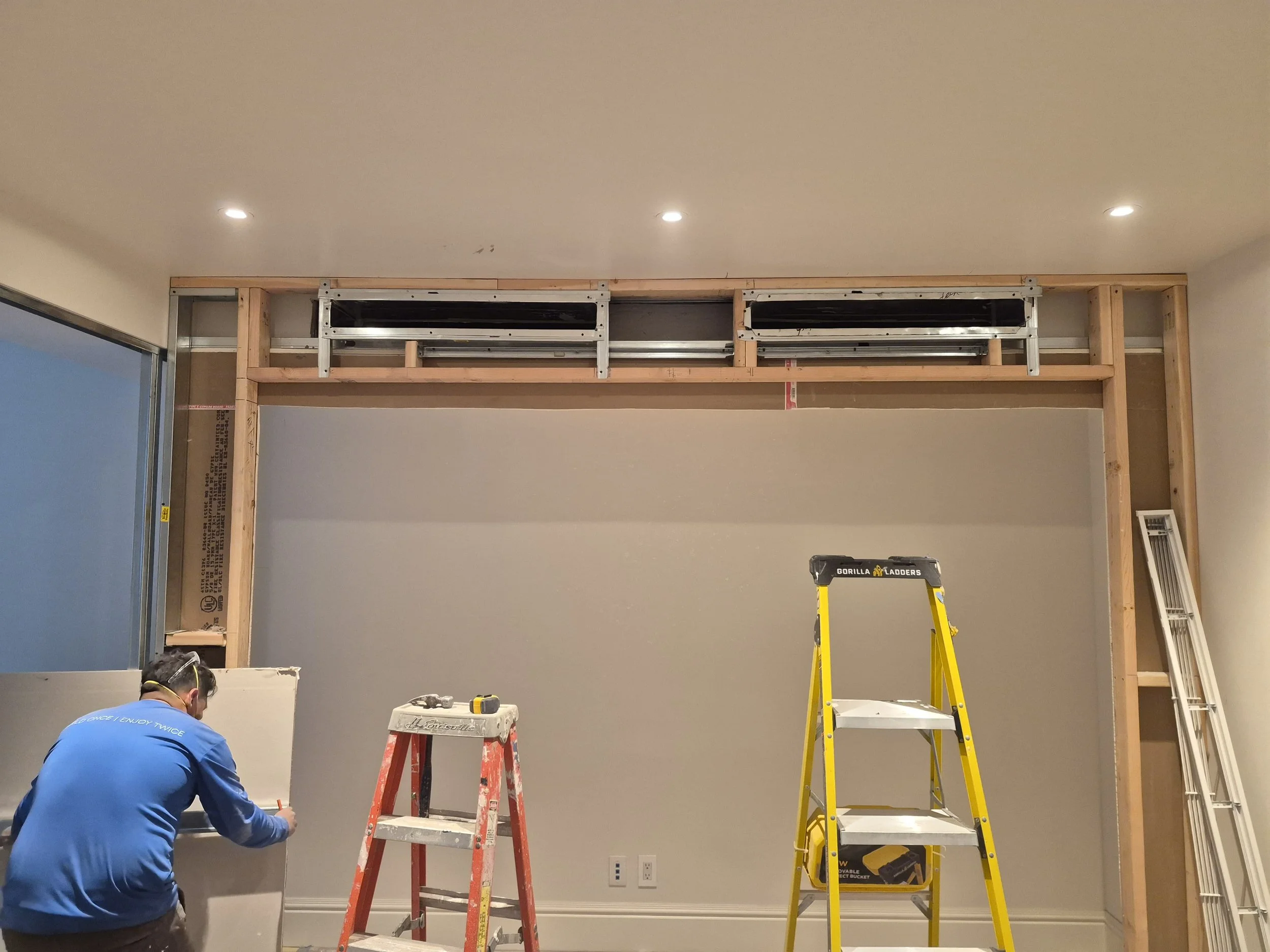 A construction worker in a blue shirt working on a wall with a ladder and scaffolding in a room under renovation.