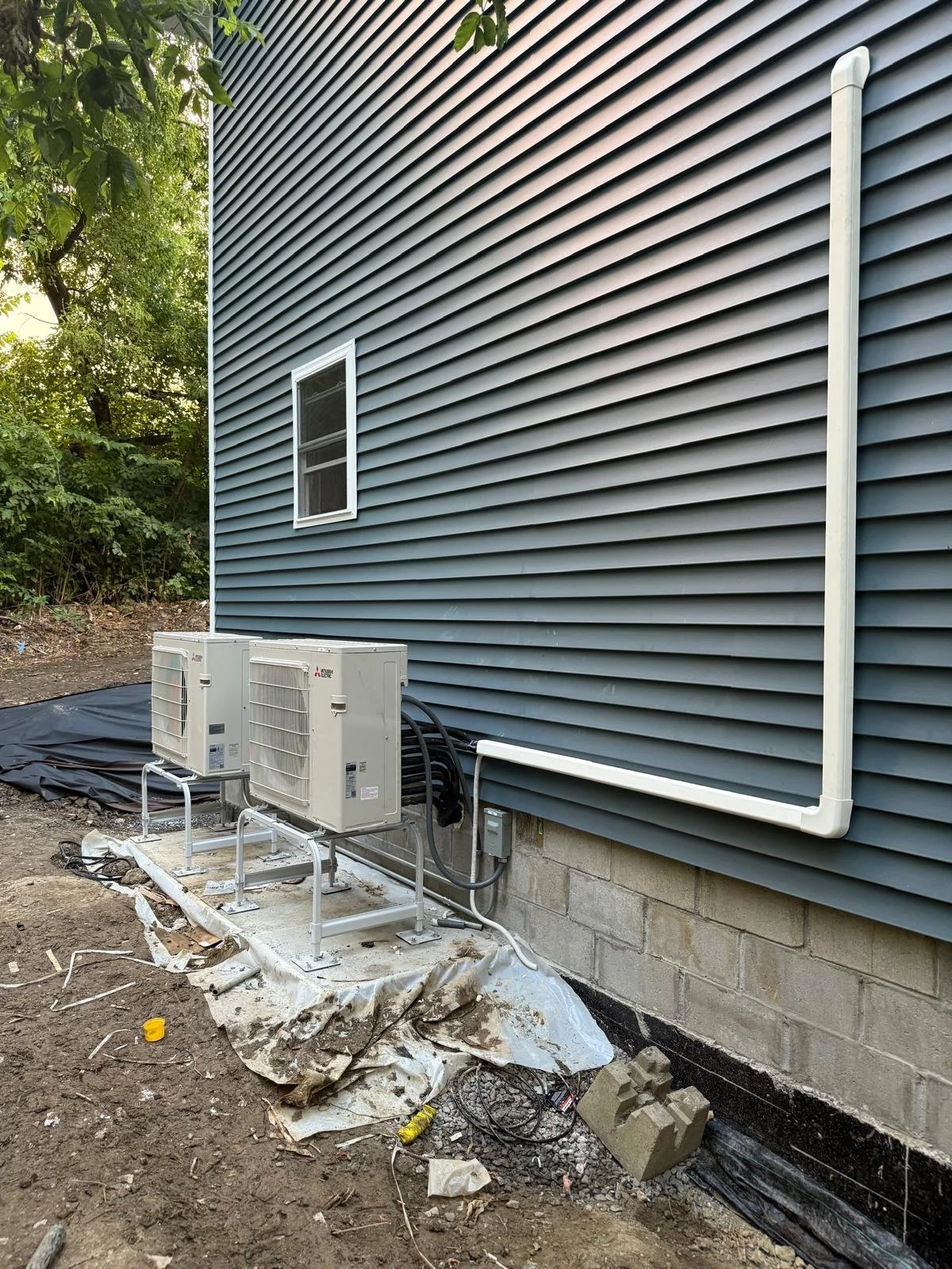 Side of a house with blue siding, showing a window, two air conditioning units, conduit pipes, and construction debris and tools on the ground.