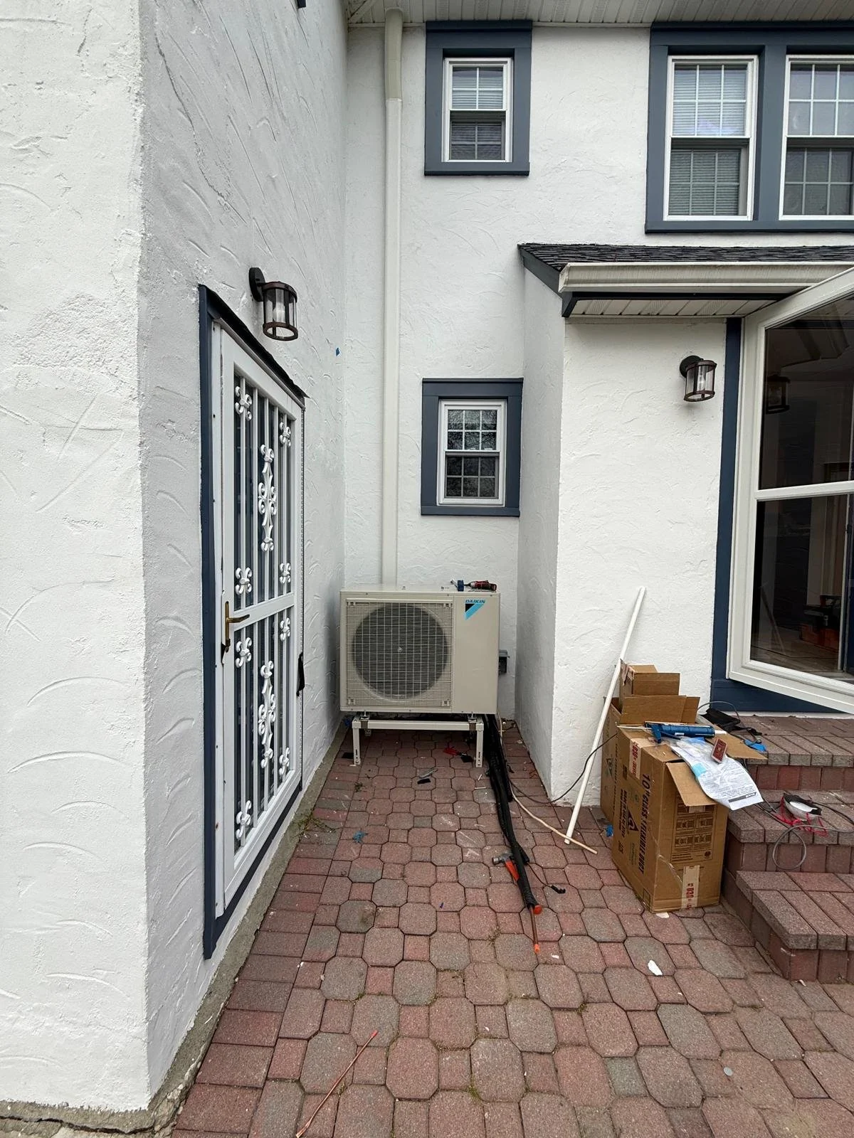 Backyard patio with paver stones, utility boxes, and a white house wall with multiple windows, outdoor wall lights, and an air conditioning unit.