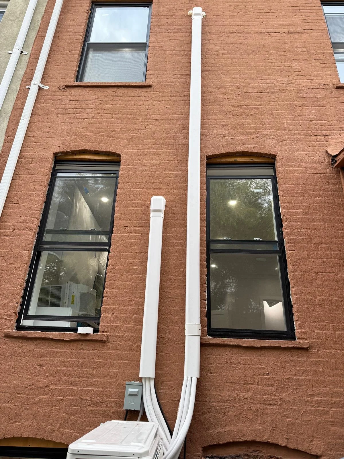 Close-up of a brick building exterior with four tall windows and two vertical white pipes running down the center, with an air conditioning unit and electrical box below.