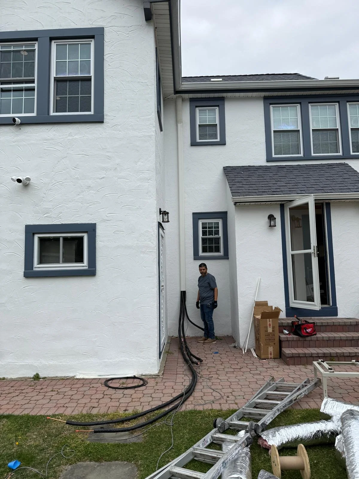 A man standing outside a white house with blue window frames, during construction or renovation, with tools and equipment on a brick patio and green lawn.