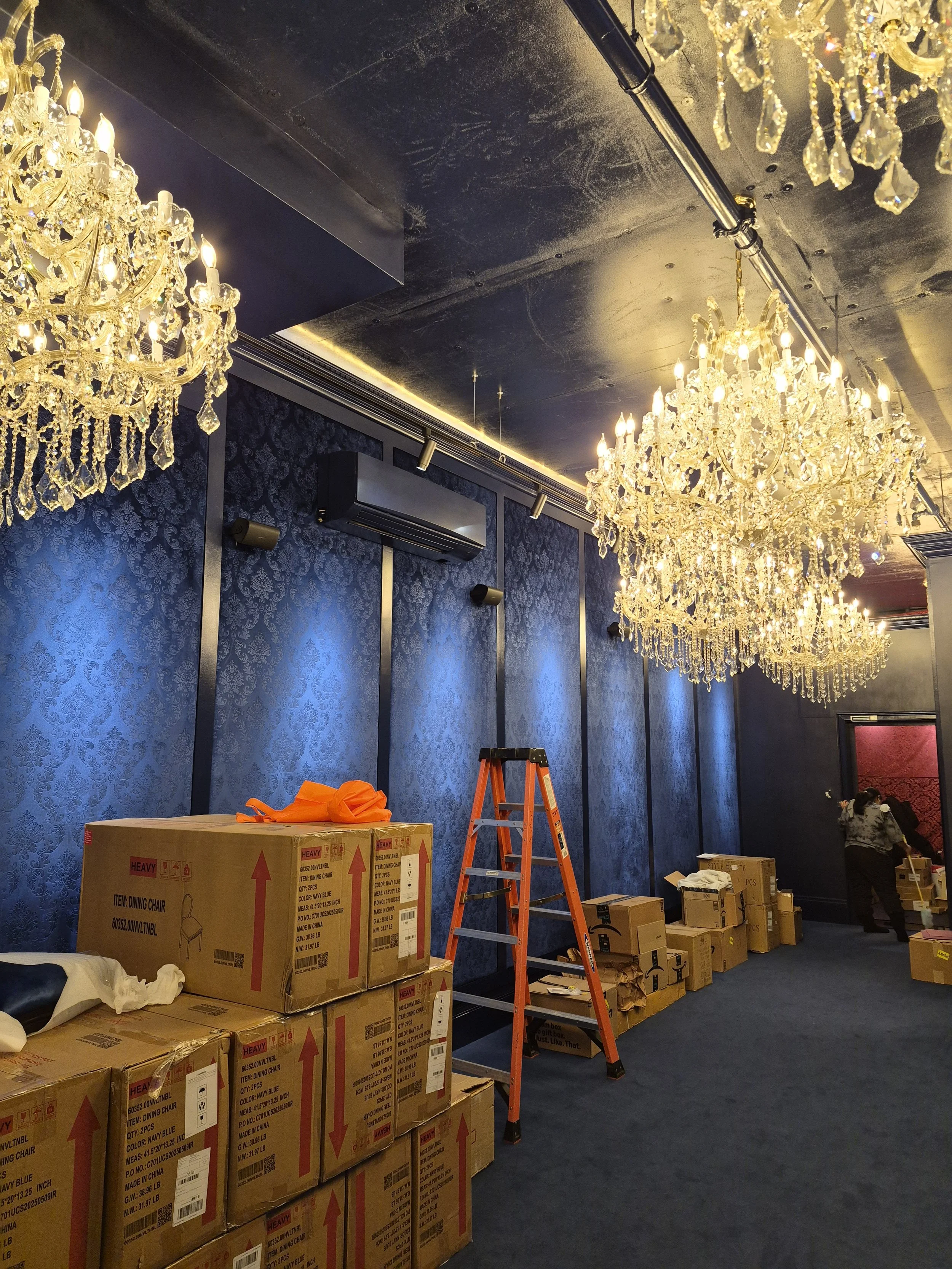 A room with blue patterned wallpaper and a dark ceiling adorned with large, ornate chandeliers. There are cardboard boxes labeled 'dining chairs' and some orange fabric or clothing on top of the boxes. An orange ladder is also present in the room, and a person is seen in the background near the wall.