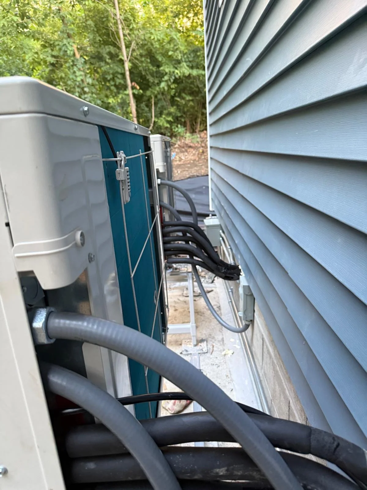 Close-up of an outdoor air conditioning unit installed against a house with gray vinyl siding, with trees and greenery in the background.