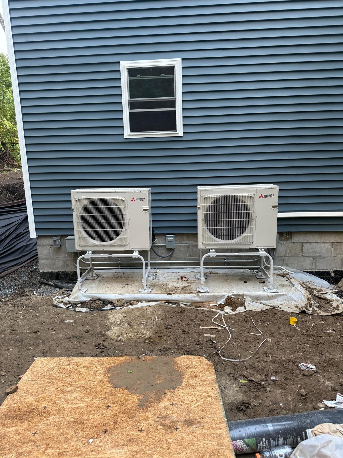 Exterior view of a house with blue vinyl siding, a small window, and two installed air conditioning units on the ground in front, with construction materials and dirt surrounding them.