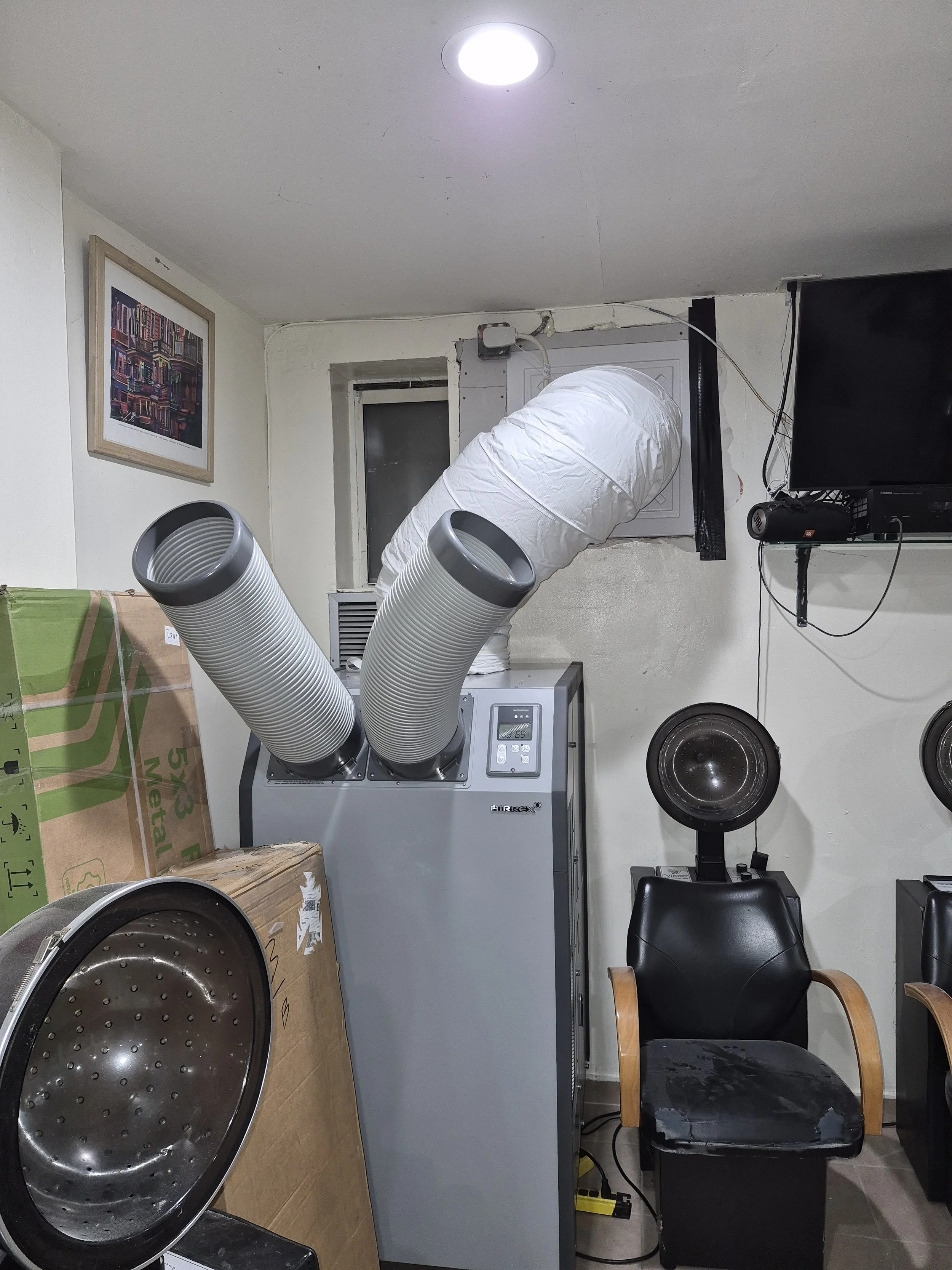 A laundry room with a large steel drum, a gray machine with two large white hoses, a black chair, a wall-mounted television, and a framed picture on the wall.