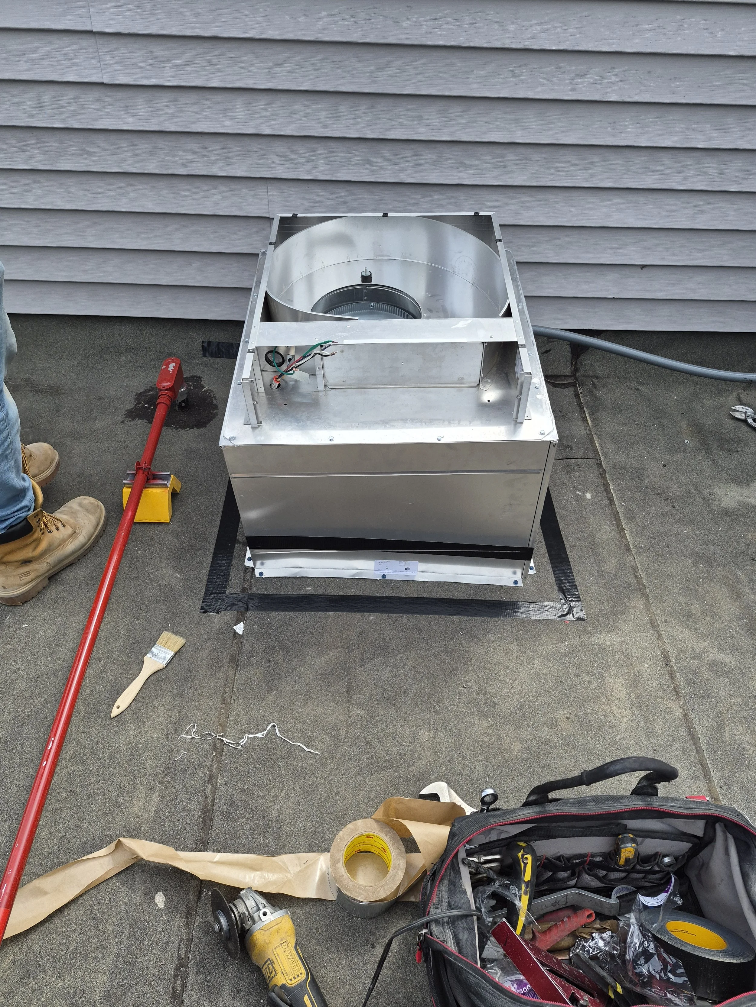 A metal outdoor air conditioning unit or HVAC system with parts and tools around it, including a yellow power drill, a paintbrush, a roll of tape, and a toolbox with various tools.