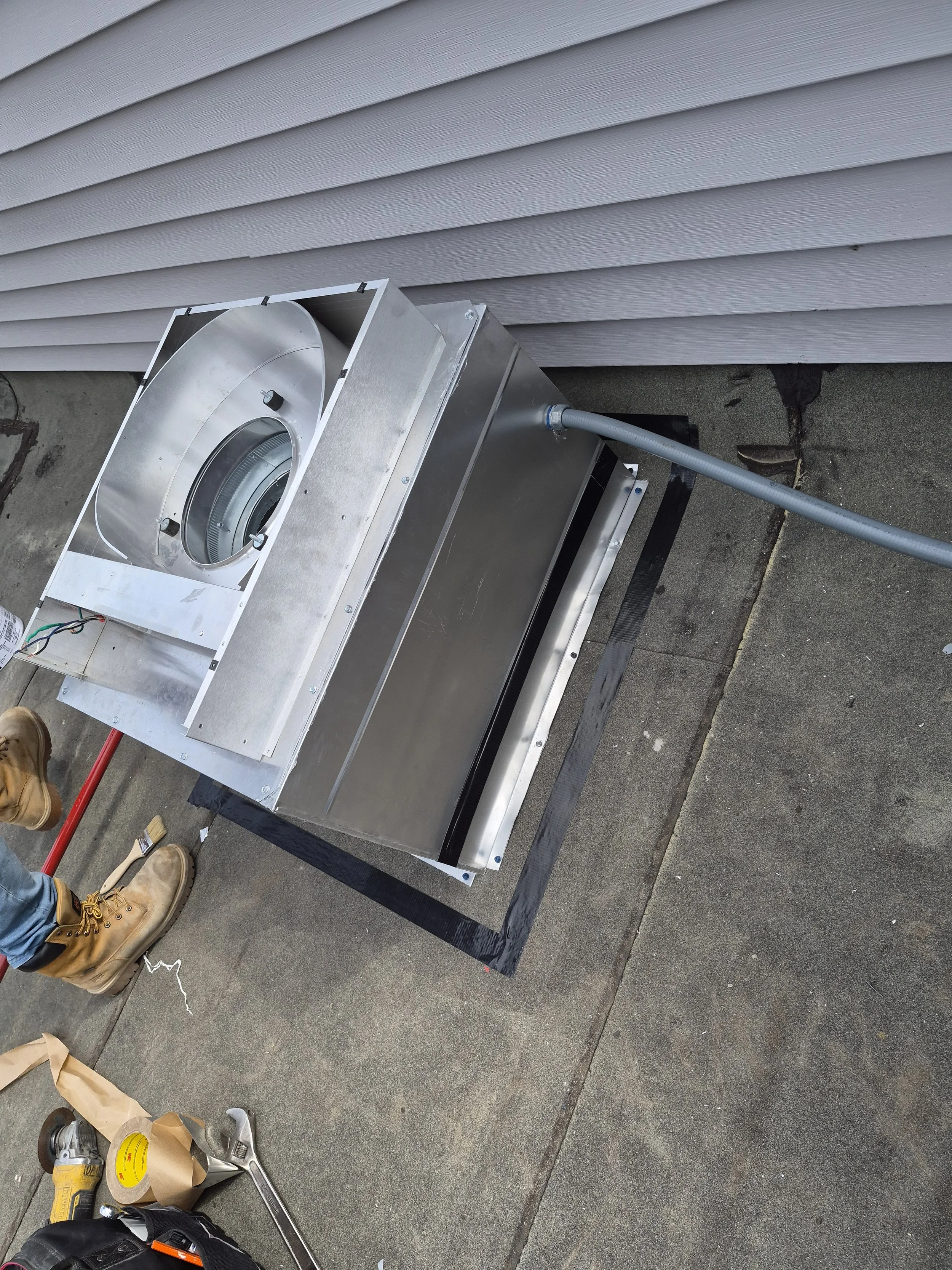 Metal HVAC or ventilation ductwork with a large circular opening, installed on a concrete surface next to a white house wall. Work tools and supplies, including a tape measure, a hammer, a wrench, and a paper bag, are on the ground nearby.